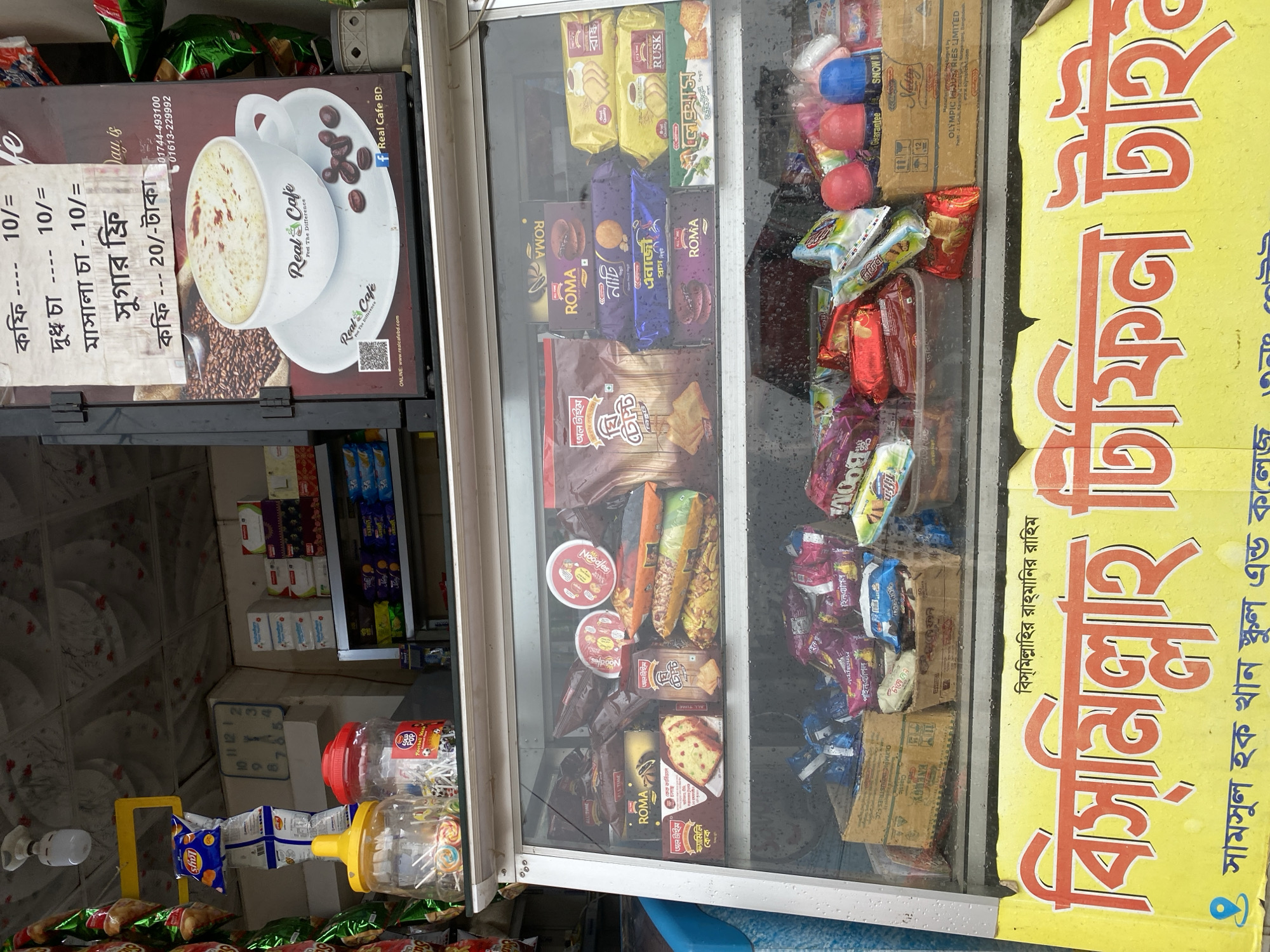 A street-side vending stall in Dhaka, Bangladesh. The stall features a glass-fronted display case containing an assortment of packaged snacks, biscuits, and candies. Above the display case is a sign advertising "Real Cafe" with coffee prices, indicating the sale of hot beverages. The items in the display case include brands like Roma, Olympic, and All Time. The stall also features a yellow sign with text in Bengali, advertising "Bismillah Tiffin Time," suggesting the sale of snack items, especially for schoolchildren. Below the display case are plastic jars containing candies and other snacks, and there are bags of chips hanging at the bottom. The location is near the "Samsul Hoque Khan School and College."