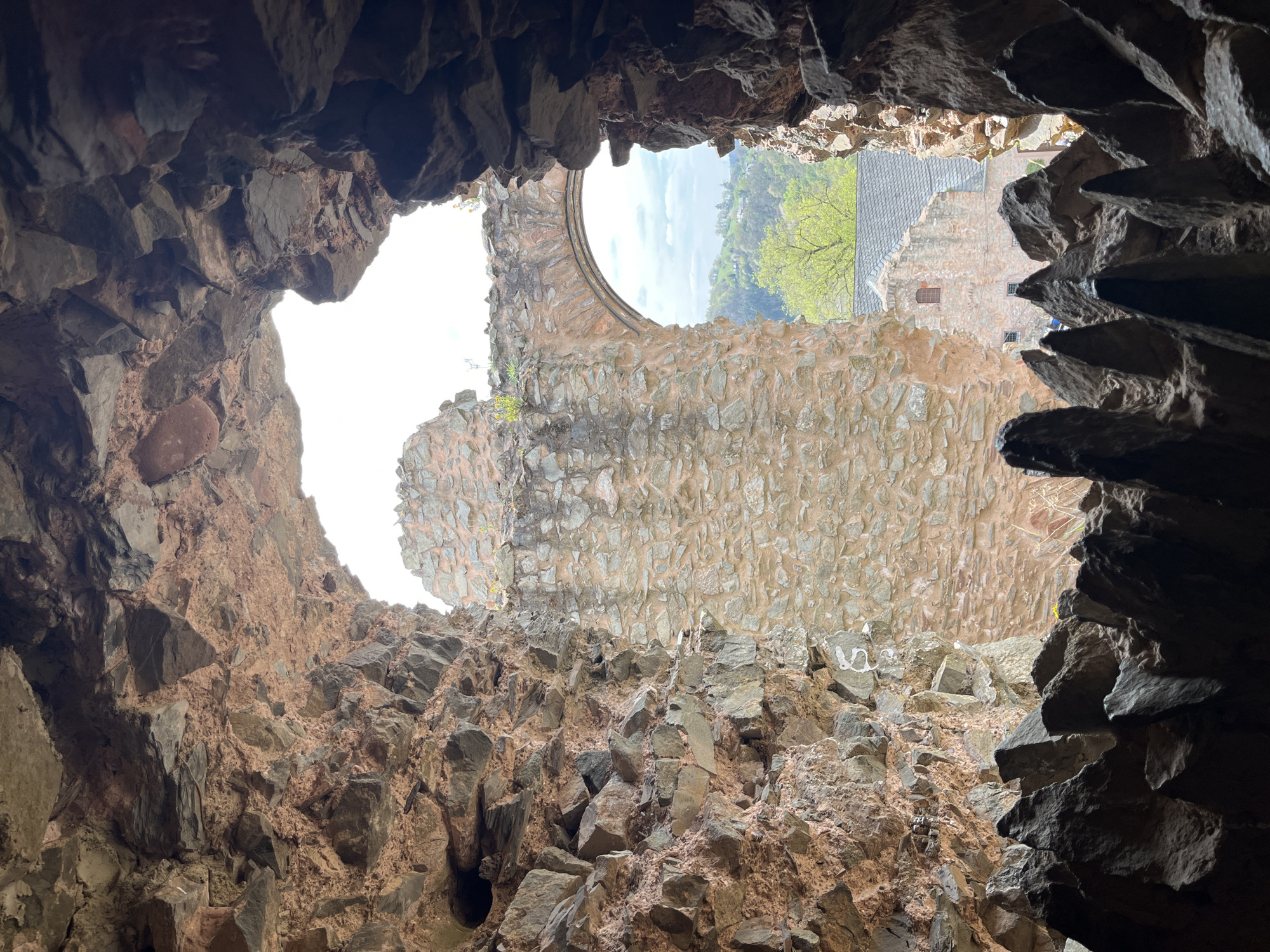 The image shows an upward view from inside the ruins of Saarburg Castle in Saarburg, Germany. The frame is defined by the jagged, rough edges of the stone interior structure. A large, irregular opening to the sky sits at the top left, while another opening revealing parts of the town and landscape is visible through the arch of another wall to the right. The stonework of the castle is made of gray and beige stone and mortar. Beyond the ruins, some buildings with gray roofs are visible, along with trees and a bit of sky.