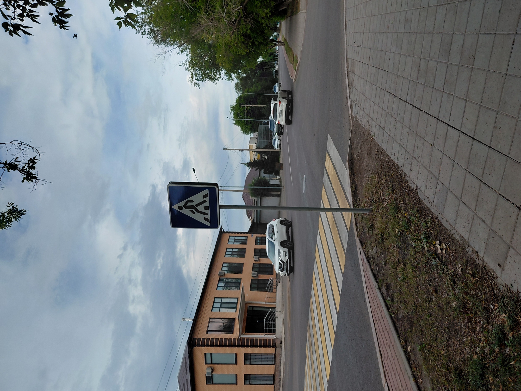 A pedestrian crossing sign is visible on a pole on the right side of the image. A white SUV is driving on the road in front of a tan brick building. The building has several windows and an entrance with railings. The sky is cloudy. The road has yellow and white stripes for a pedestrian crossing. There is a sidewalk on the right side of the image.