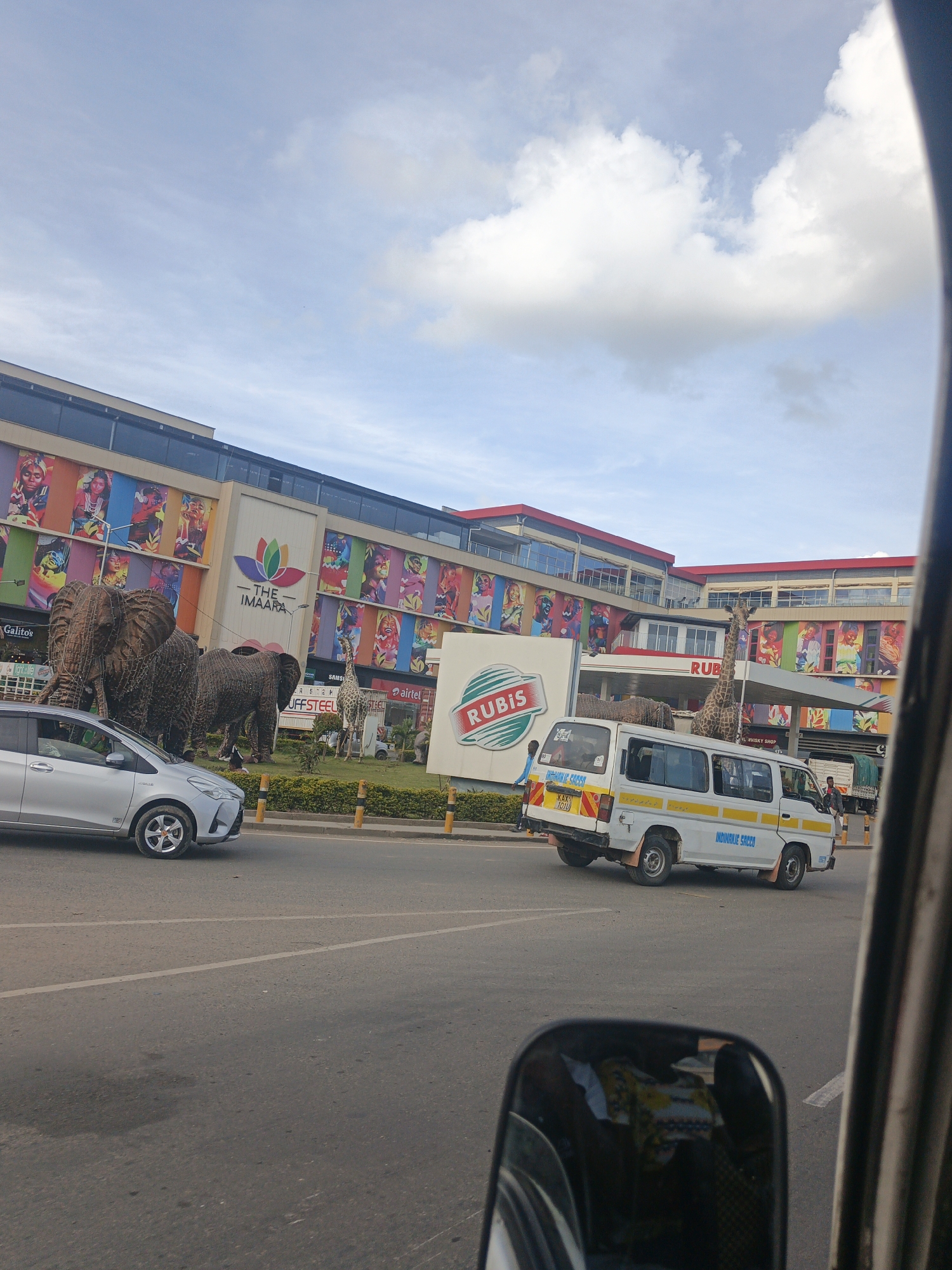 The image shows a street view in Nairobi, Kenya, with "THE IMAARA" shopping mall prominently displayed in the background. Wire sculpture elephants and a giraffe are in front of the mall, near a Rubis petrol station. A silver car and a white van with "INDIMANJE SACCO" written on it are on the road. A person is visible near the van. The photo appears to be taken from inside another vehicle, with part of the vehicle's interior visible in the foreground, including the side mirror. The sky is partly cloudy.