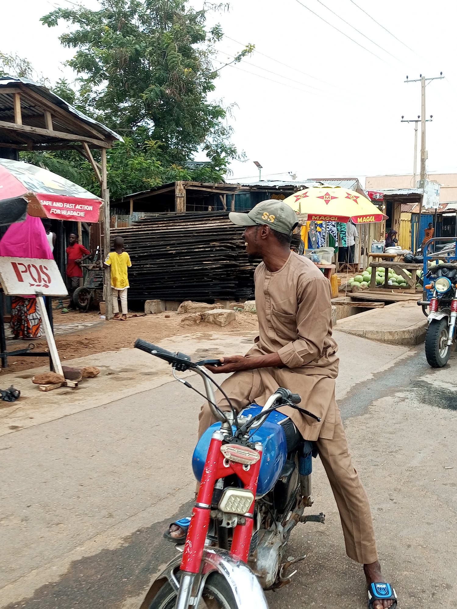 A man wearing a tan-colored traditional outfit and a green baseball cap is seated on a blue and red motorcycle. He is looking to his right. The motorcycle has a silver handlebar and a visible front tire. The man's feet are clad in blue and black sandals. The scene appears to be outdoors in a market setting in Gombe, Nigeria, indicated by the presence of market stalls, umbrellas, and stacked wooden planks. In the background, a red and white umbrella with the text "SAFE AND FAST ACTION FOR HEADACHE" is visible. Another yellow umbrella with the "MAGGI" logo is also present. Several watermelons are displayed on a wooden structure to the right. Power lines are visible in the upper portion of the image. The ground is paved with asphalt.