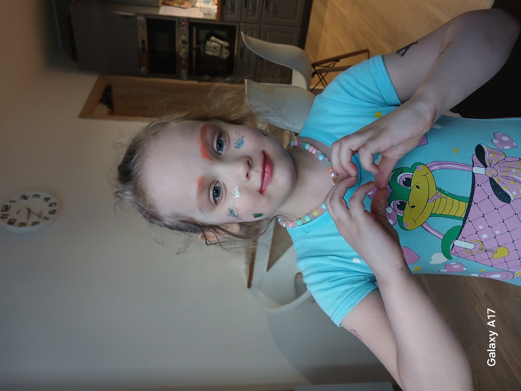A young child is shown indoors, likely in a residence given the presence of kitchen appliances and furniture. The child has light skin, fair hair, and blue eyes, with makeup or glitter applied to the face around the eyes. The child is wearing a blue T-shirt with a cartoon design featuring a green crocodile or dragon and a pink checkered handbag. The child's hands are positioned near the face, as if making a gesture, and a beaded necklace is visible around the neck. The location is unknown so information about Yekaterinburg, Russia, cannot be added. A clock on the wall indicates a time of around 4:53, though the AM/PM status is undetermined. The room is somewhat illuminated with natural and artificial light. The camera used to take the photo appears to be a Samsung Galaxy A17, as indicated by the text in the lower-right corner of the image.