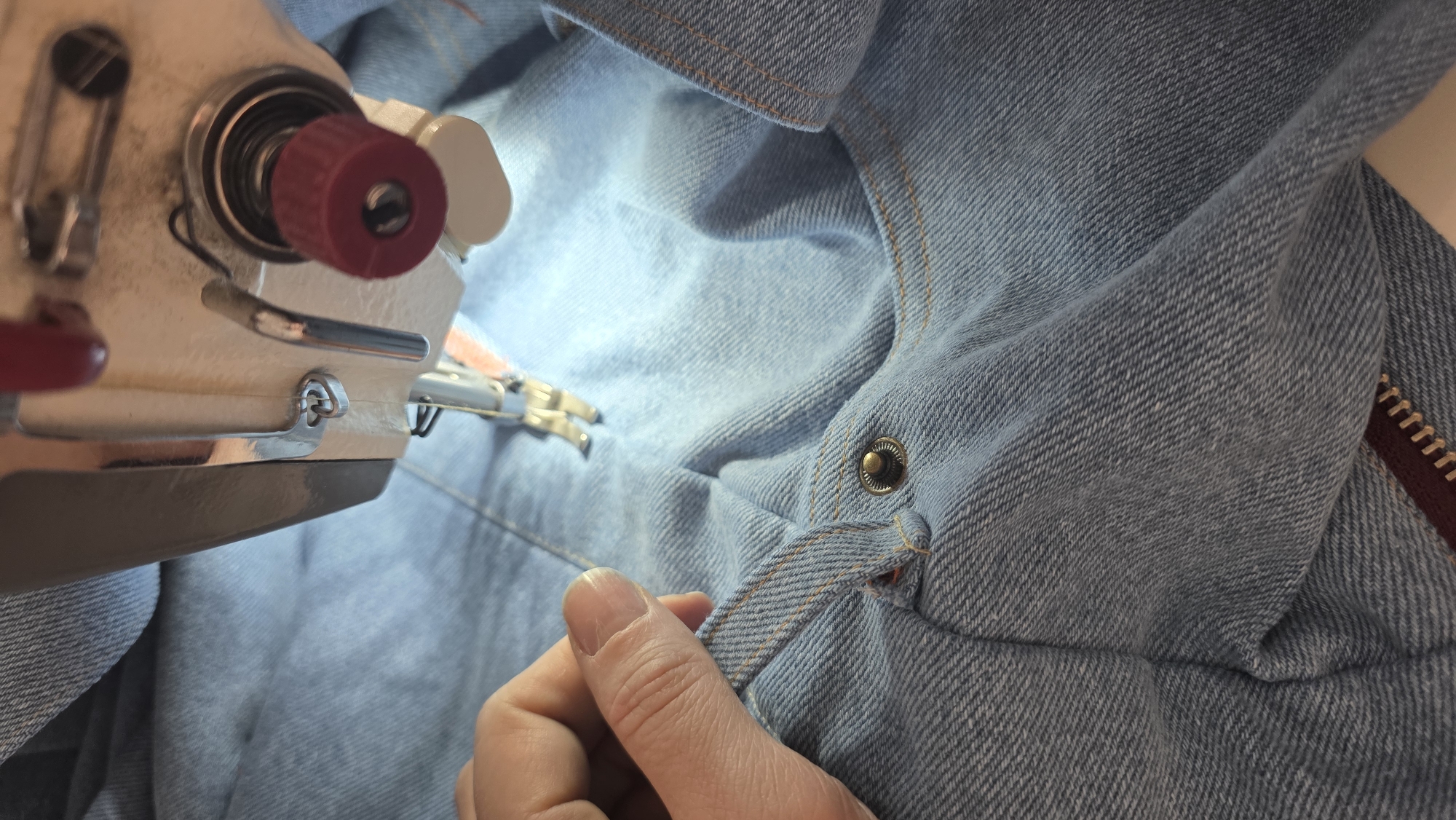 A close-up shot depicts a person using a sewing machine to mend a light blue denim garment. The sewing machine, visible on the left, is light-colored with red and silver accents. A finger of the person’s hand is visible holding a denim loop. The denim garment also features a snap and a zipper on the right side of the frame. The setting of the image cannot be determined from the visual information provided.