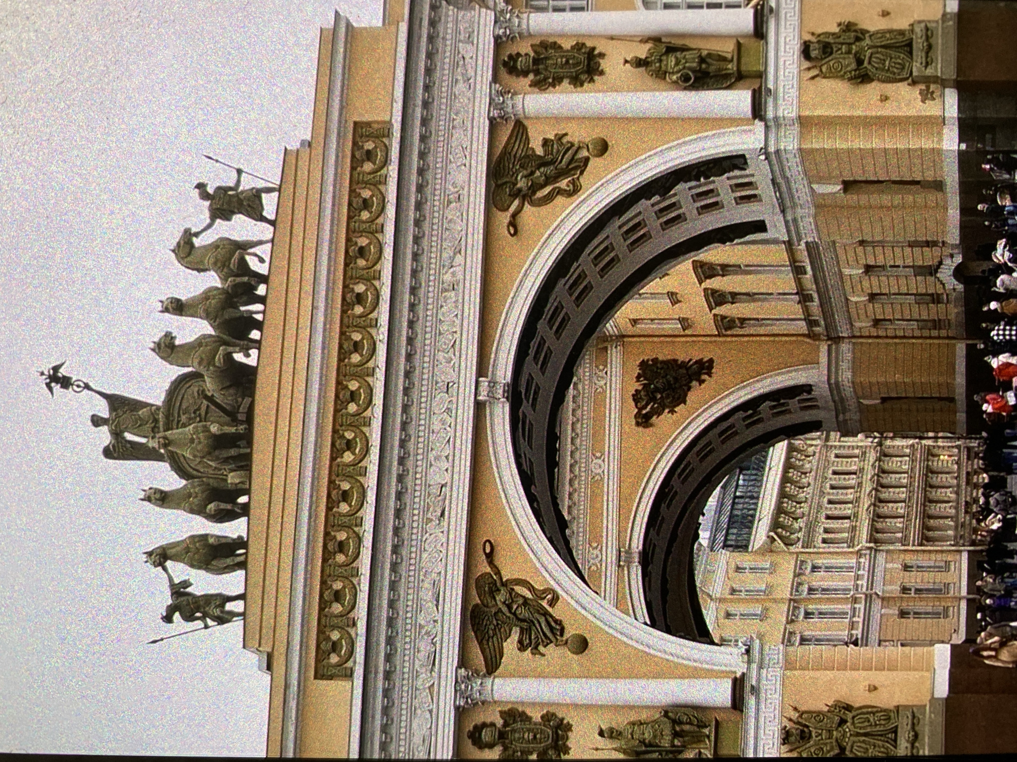 The image shows the Triumphal Arch in Palace Square, Saint Petersburg, Russia, angled from left to right. The arch features a triumphal chariot drawn by six horses atop. Below are intricate architectural details including decorative carvings, statues, and arched passageways. A crowd of people is visible to the right of the arch, and other buildings can be seen through the archways. The sky is overcast.