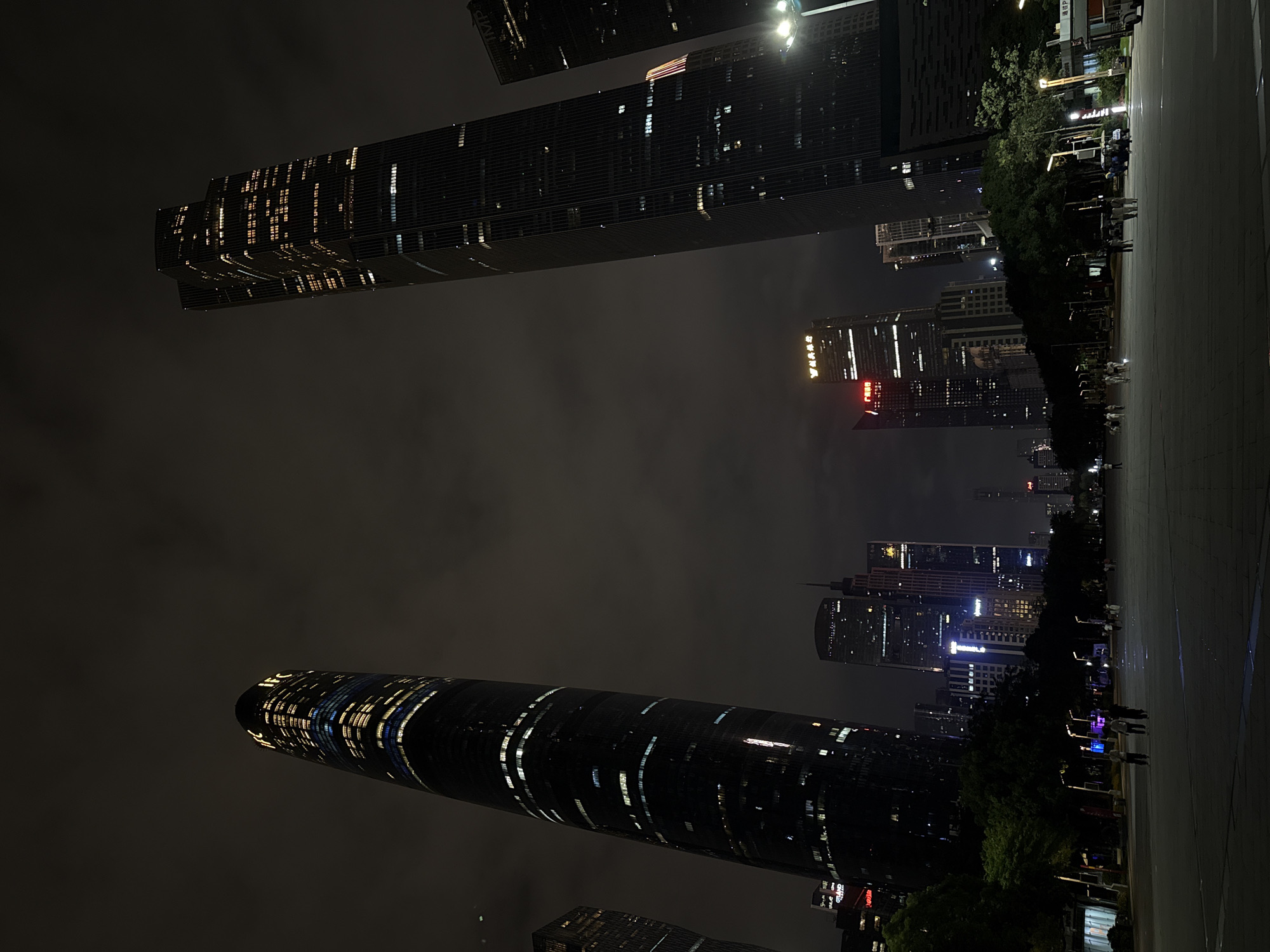 This nighttime photo shows the skyline of Tianhe District, China, with several skyscrapers illuminated. The tallest building appears cylindrical and stretches diagonally across the lower left. Another tall, rectangular building is visible near the top. Between the buildings, there is a dark, cloudy sky. On the right is a dark, flat walkway with dark trees and street lights. People are seen walking along the walkway. The image has a slightly tilted perspective.