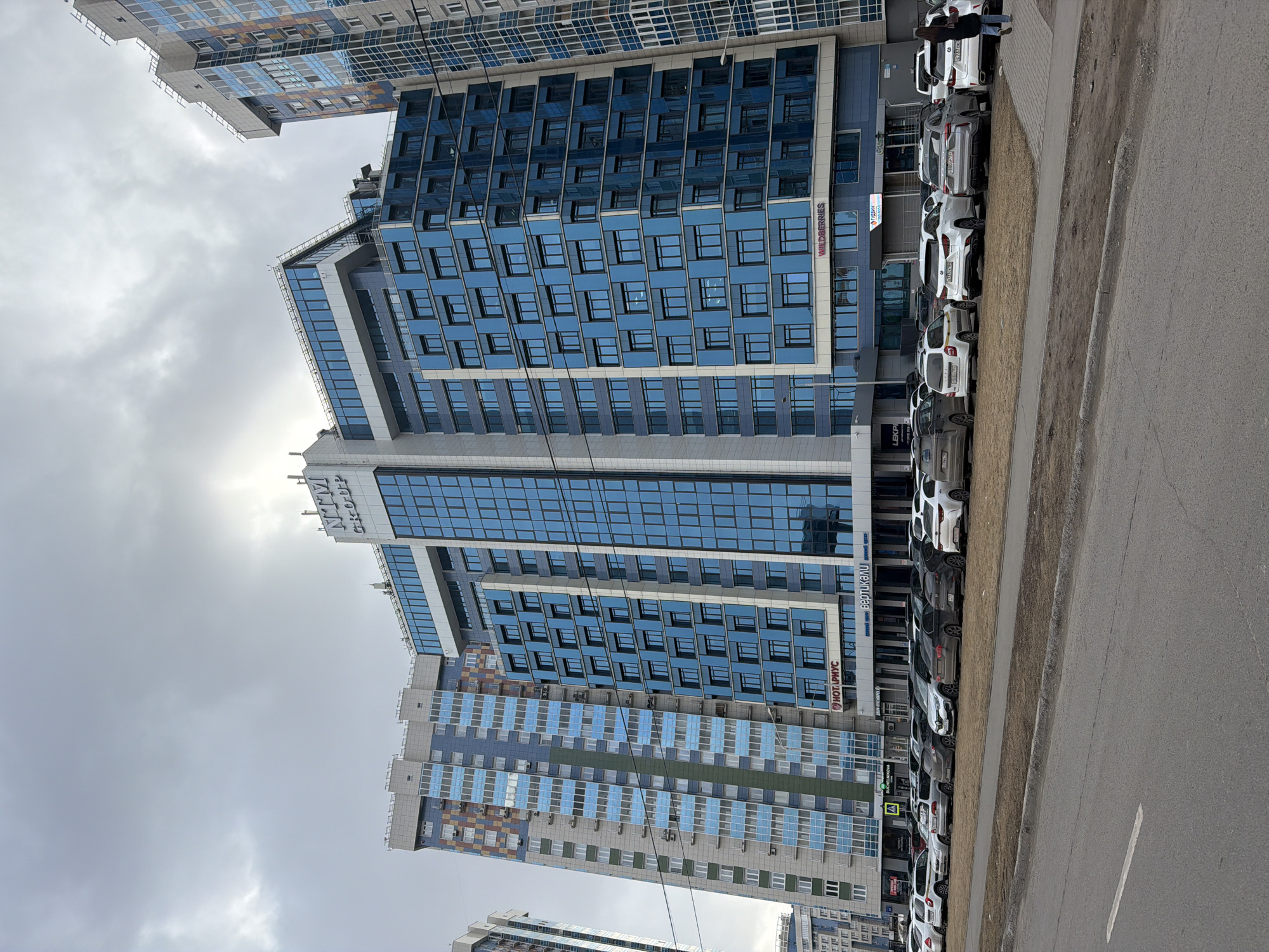 This urban street scene, set in Krasnoyarsk, Russia, captures a view dominated by modern high-rise buildings under a bright, overcast sky. The prominent subject is a tall, multi-story building featuring a contemporary blue glass and grey panel facade, reflecting the cloudy daylight. This building appears to be mixed-use, housing businesses and potentially residential units. Several signs are visible on its exterior, including "WILDERRIES" (likely Wildberries, a major online retailer), "ПУРПУР" (Purpur), "НОТАРИУС" (Notary), and "ЛЕКАРС" (Lekar, suggesting a pharmacy). The "NTT GROUP" logo is displayed at the top of its tallest section.
To the left and in the background, other residential or commercial high-rises are visible, showcasing diverse architectural styles with facades in shades of blue, grey, and white, some featuring distinct checkerboard patterns.
In the foreground and running along the right side of the image, a paved road parallels a sidewalk. A narrow strip of dirt and sparse grass separates the roadway from the pedestrian path. A long queue of cars, primarily sedans and SUVs in white, black, and grey, are parked curb-side along the buildings. One individual is clearly visible standing on the sidewalk near the parked vehicles, looking towards the buildings or street. Another person is partially visible near the rooftop of the main blue building.
The time of day is daytime, characterized by a bright, diffused light from the cloud-covered sky, indicating an overcast weather condition. The scene depicts a bustling, modern urban environment with commercial activity and residential presence, typical of a city district. All visible text is in Russian, consistent with the location in Russia.