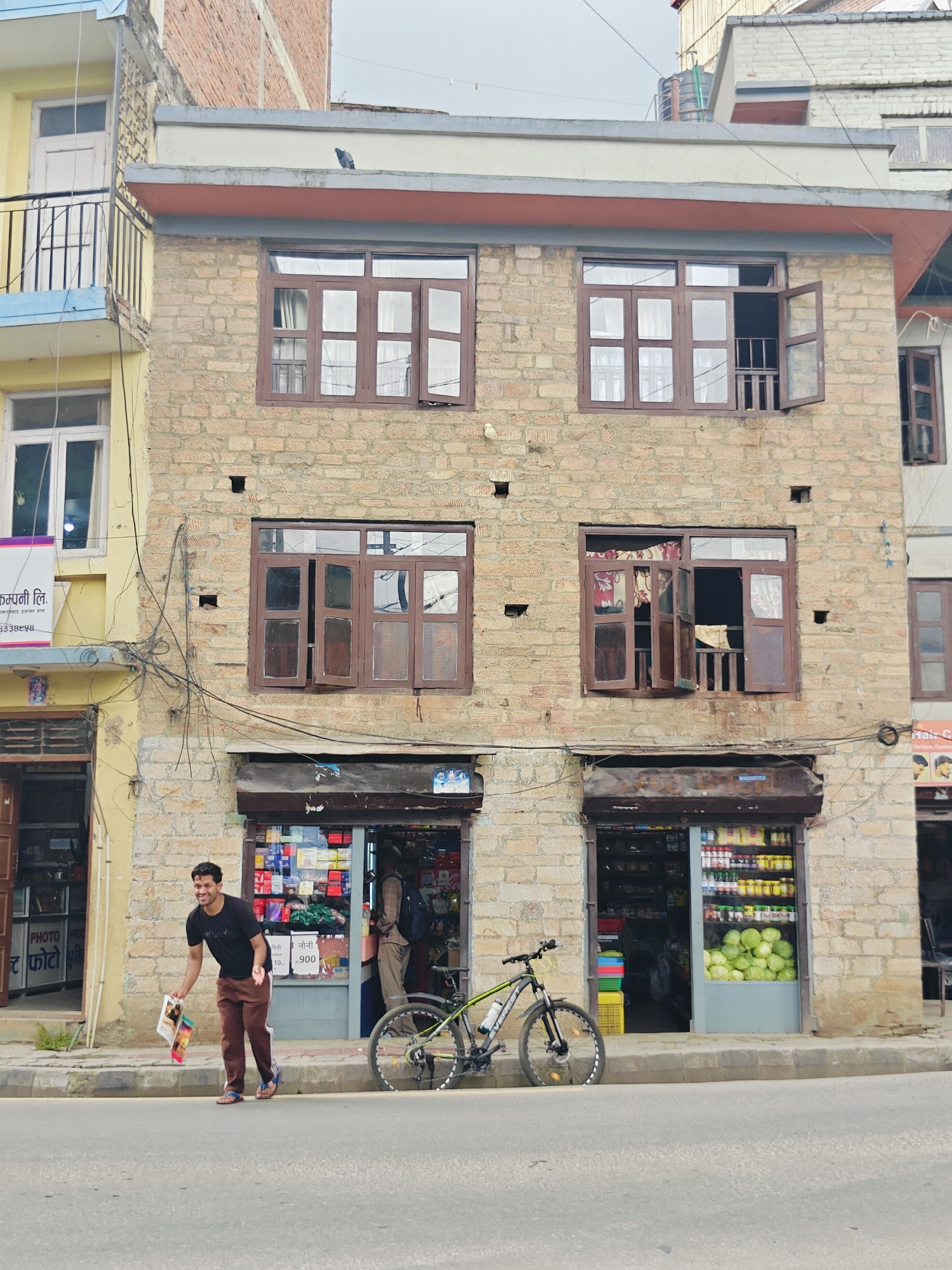 In Kirtipur, Nepal, a man in a black t-shirt and brown pants stands on a sidewalk in front of a two-story brick building. He is smiling and holding a few colorful papers. To his right, a bicycle with a water bottle is leaning against the building. The building has multiple windows, some open, some closed, and two storefronts on the ground floor. One storefront displays various goods, including jars of food and a large pile of green cabbage. The other storefront, partially visible to the left, has a "PHOTO PRO" sign. The sidewalk is concrete, and the street in front is paved. Overhead, electrical wires crisscross the facade of the building. A pigeon is perched on the roofline of the building. The sky is overcast.