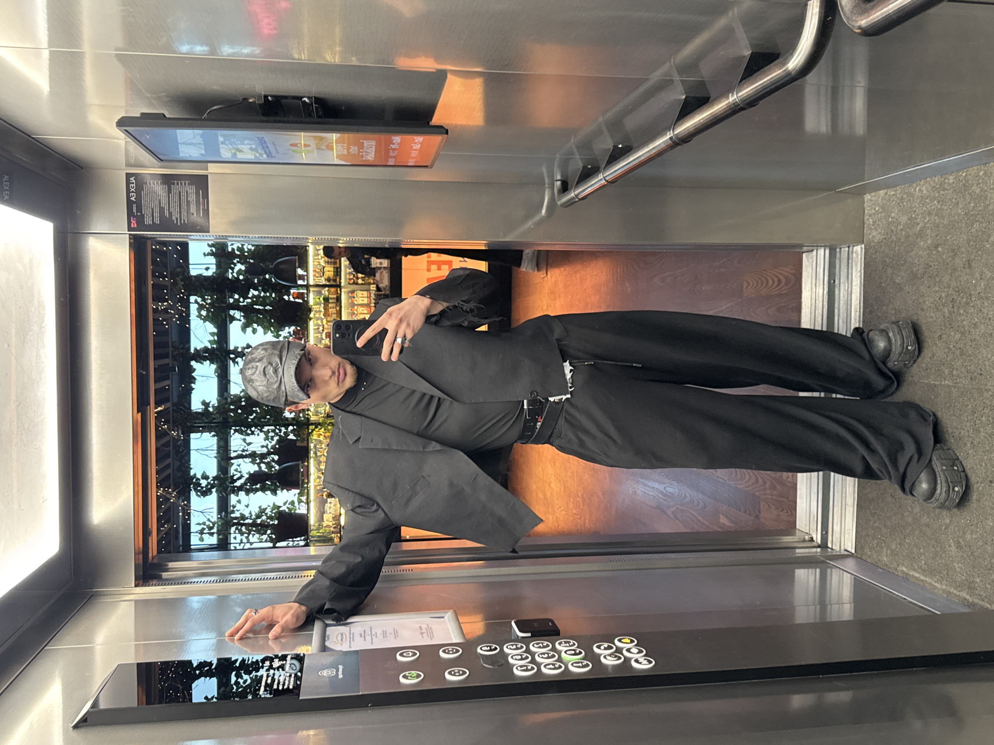 A person is taking a selfie in an elevator. The individual is wearing black clothing, including a blazer, pants, shoes, and a hat. The elevator has stainless steel walls and a control panel with buttons. A digital display screen shows a building exterior with trees and other features visible. The elevator opens to a building interior with a bar or restaurant visible through the opening. Based on the style of the interior and exterior view, this is most likely at The Mall in Sofia, Bulgaria.