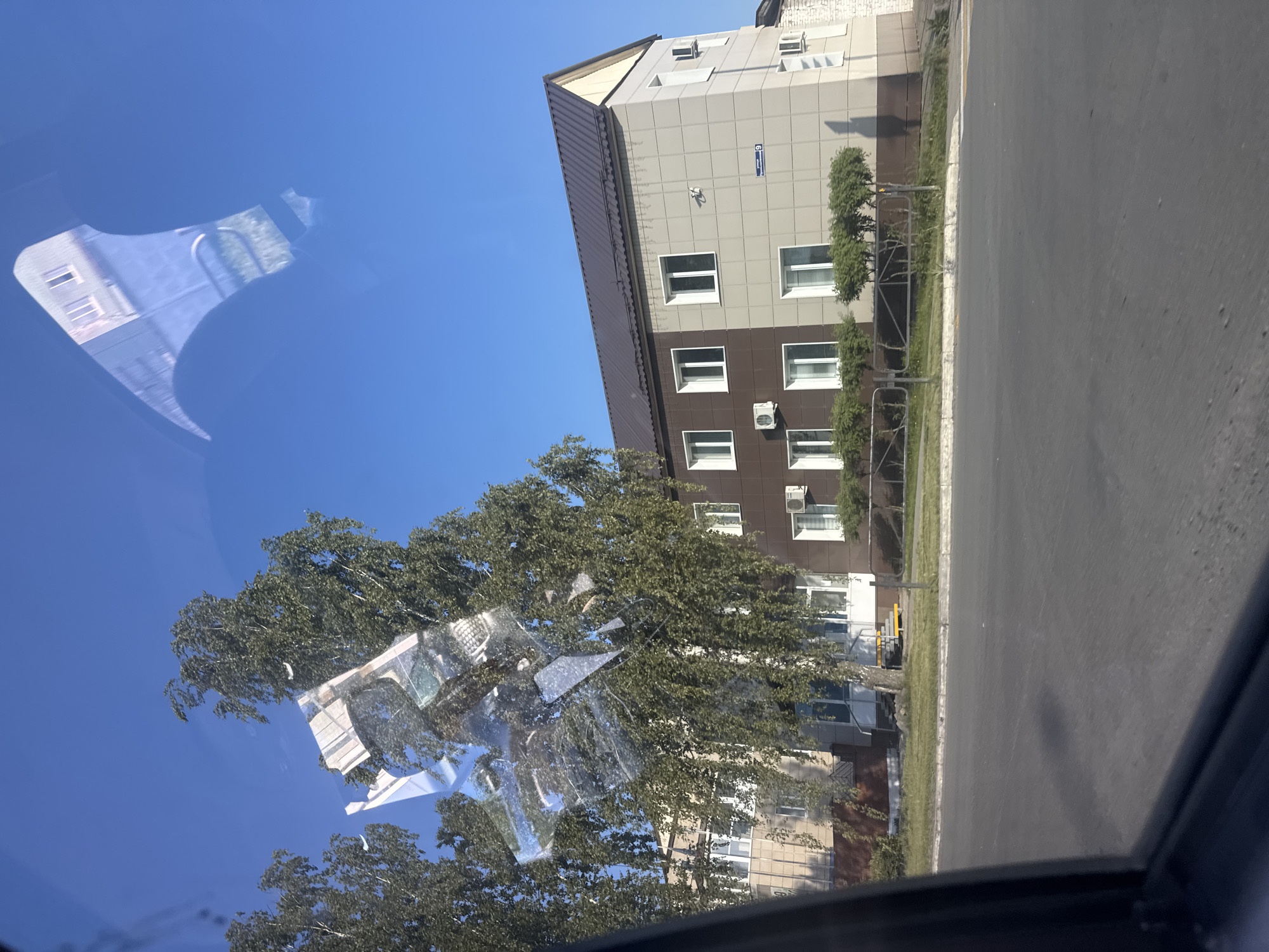 The image depicts a street scene, likely taken from inside a vehicle, in Chaikovsky, Russia. In the background is a multi-story building, partially obscured by trees. The building has a gray and brown facade, with white-framed windows and visible air conditioning units. A street sign is mounted on the building. The foreground includes a car window with reflections of trees and the building, distorting the view of the sky. A paved road extends to the right of the building.
