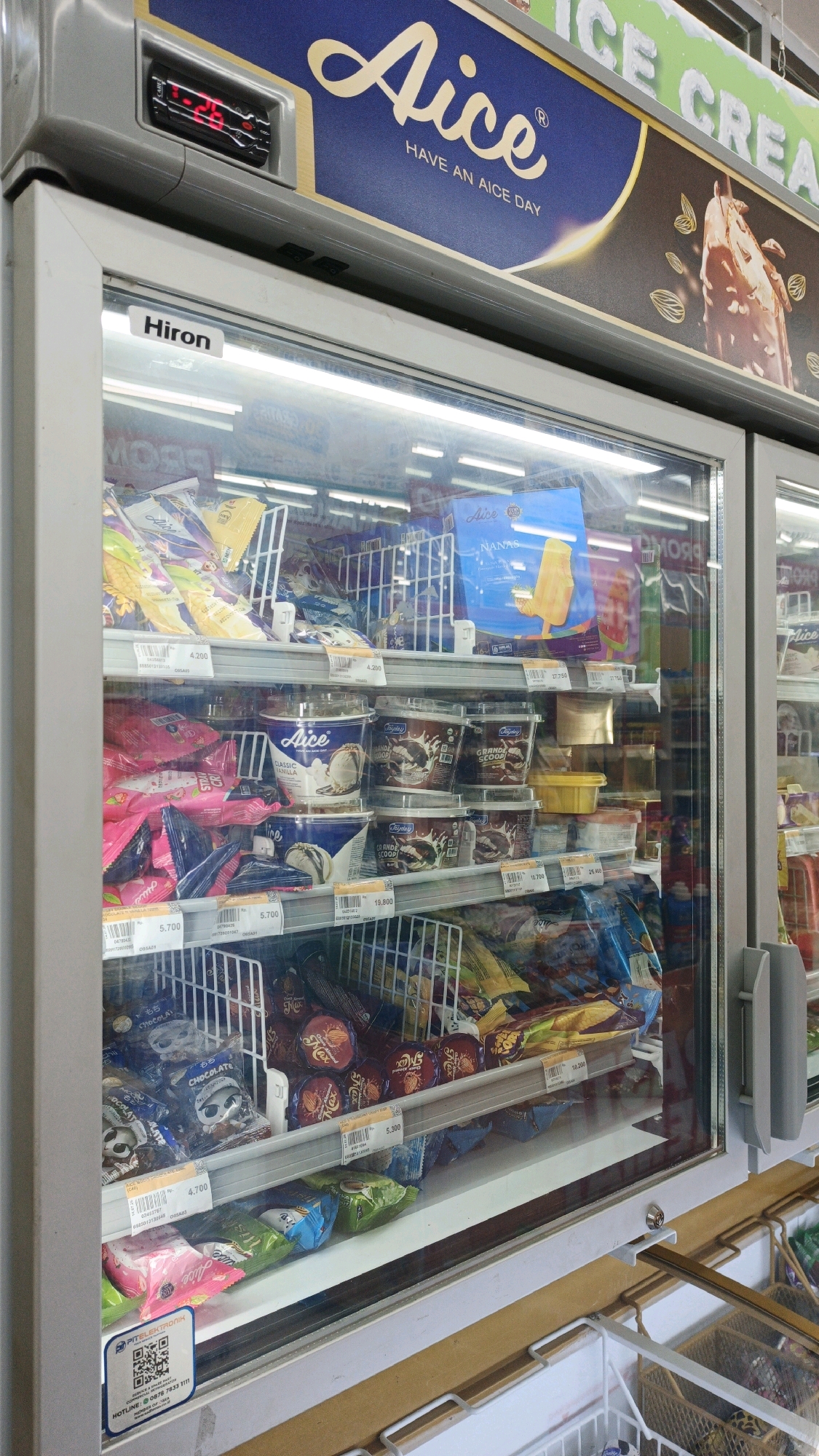 The image shows a freezer display case filled with various ice cream products. The freezer has a glass door, allowing a clear view of the items inside. Above the freezer door, a large sign displays the brand "Aice" with the slogan "HAVE AN AICE DAY". There is also a digital display showing "26" degrees Celsius, indicating the temperature inside the freezer. The shelves are stocked with individually wrapped ice cream bars, tubs of ice cream, and other frozen treats. Some of the packaging features colorful designs and brand logos, including "Aice", "Nanas", "Classic Vanilla", and "Grande Scoop". Price tags are visible on some of the products. The overall environment appears to be a retail setting, likely a convenience store or a supermarket, in Parigi, Indonesia.