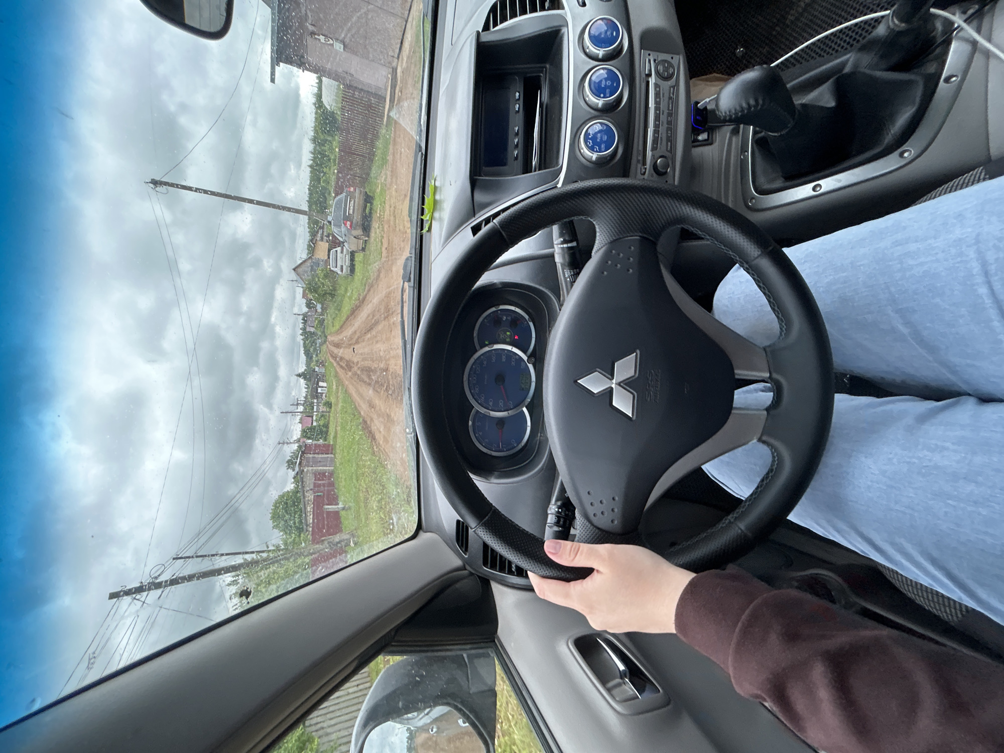 The image offers a first-person view from the driver's seat of a car, looking out onto a rural landscape. A person's right hand, wearing a brown-sleeved top, rests on a black leather-wrapped steering wheel, which prominently displays a silver Mitsubishi logo. The driver's leg, clad in light blue jeans, is visible to the right.
Inside the car, the dashboard features an instrument cluster with blue-toned gauges, a central display screen, and three circular blue-lit dials below it. A car stereo head unit and a manual gear shifter are also visible.
The exterior scene, viewed through a windshield with minor specks (possibly rain or dust), shows an unpaved dirt road stretching into the distance. The sky is overcast and cloudy, suggesting daytime. Along the road, wooden utility poles with power lines are present. In the background, several parked vehicles and various buildings, including what appear to be houses or sheds with wooden fences, line the path. One of the parked vehicles is a grey SUV, likely a Mitsubishi, given the steering wheel logo. The setting strongly suggests a village or countryside environment, specifically in Фоки, Russia, where the location is situated. The activity appears to be driving or preparing to drive. No specific emotions are discernible.