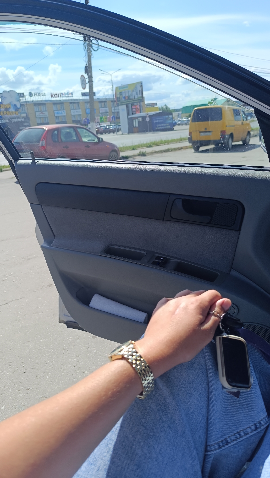 An outdoor, eye-level, medium shot captures a view from inside a vehicle looking out through the open passenger door. The focus is on the hand and forearm of an individual wearing a gold watch and blue jeans, holding car keys. The arm is positioned across the lap. In the background, across a paved road, a yellow van is parked alongside a building complex featuring businesses like "FDGA" and "Kan1-D-S". A red car is also visible in front of the buildings. The sky is blue with scattered clouds, and utility poles are present. The scene appears to be located in the city of Chaikovsky, Russia.