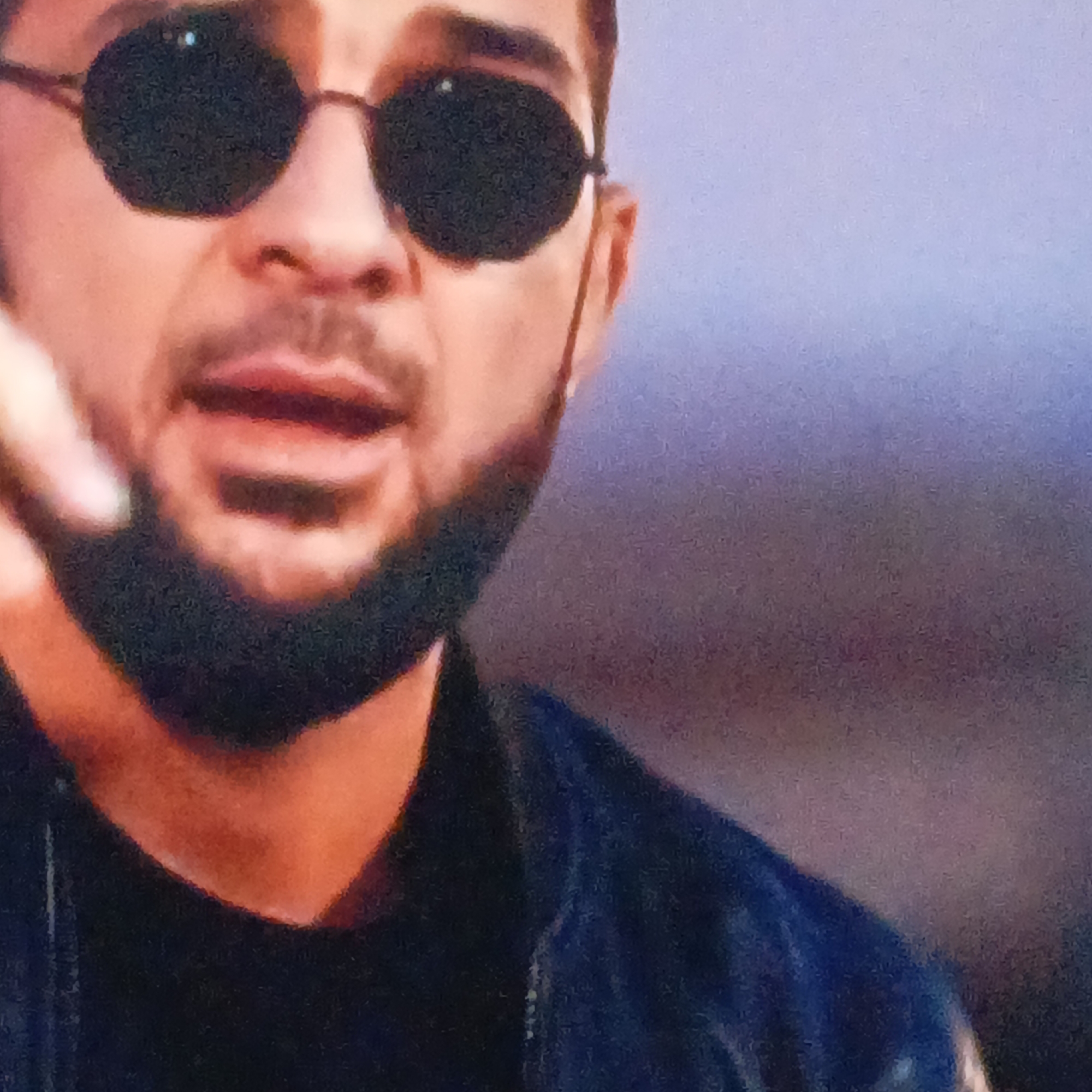 A man is captured in a mid-shot, wearing dark, round-lensed sunglasses with thin frames, which obscure his eyes. He has a dark beard and mustache, and his mouth is slightly open, suggesting he is speaking, singing, or in mid-utterance. A finger, likely from his left hand, is partially visible on the far left, appearing to gesture or point. He is dressed in a dark-colored garment, possibly a black t-shirt or shirt, layered under a dark, potentially blue, jacket or coat. The background is out of focus, consisting of muted tones of light blue and purple, with some darker, warmer hints at the bottom, creating a soft, indistinct backdrop. The overall image has a grainy texture. No specific location, time of day, or overt emotions are discernible. No text is visible in the image.