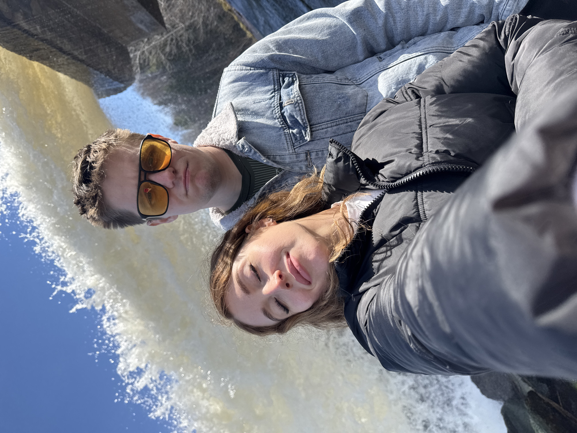 A man and a woman are posing for a selfie with a waterfall in the background. The man is wearing sunglasses and a denim jacket with a shearling collar. The woman is wearing a black puffer jacket. The sky is blue. The location is unknown, and there is no mention of the city of Noviy, Russia in the image.