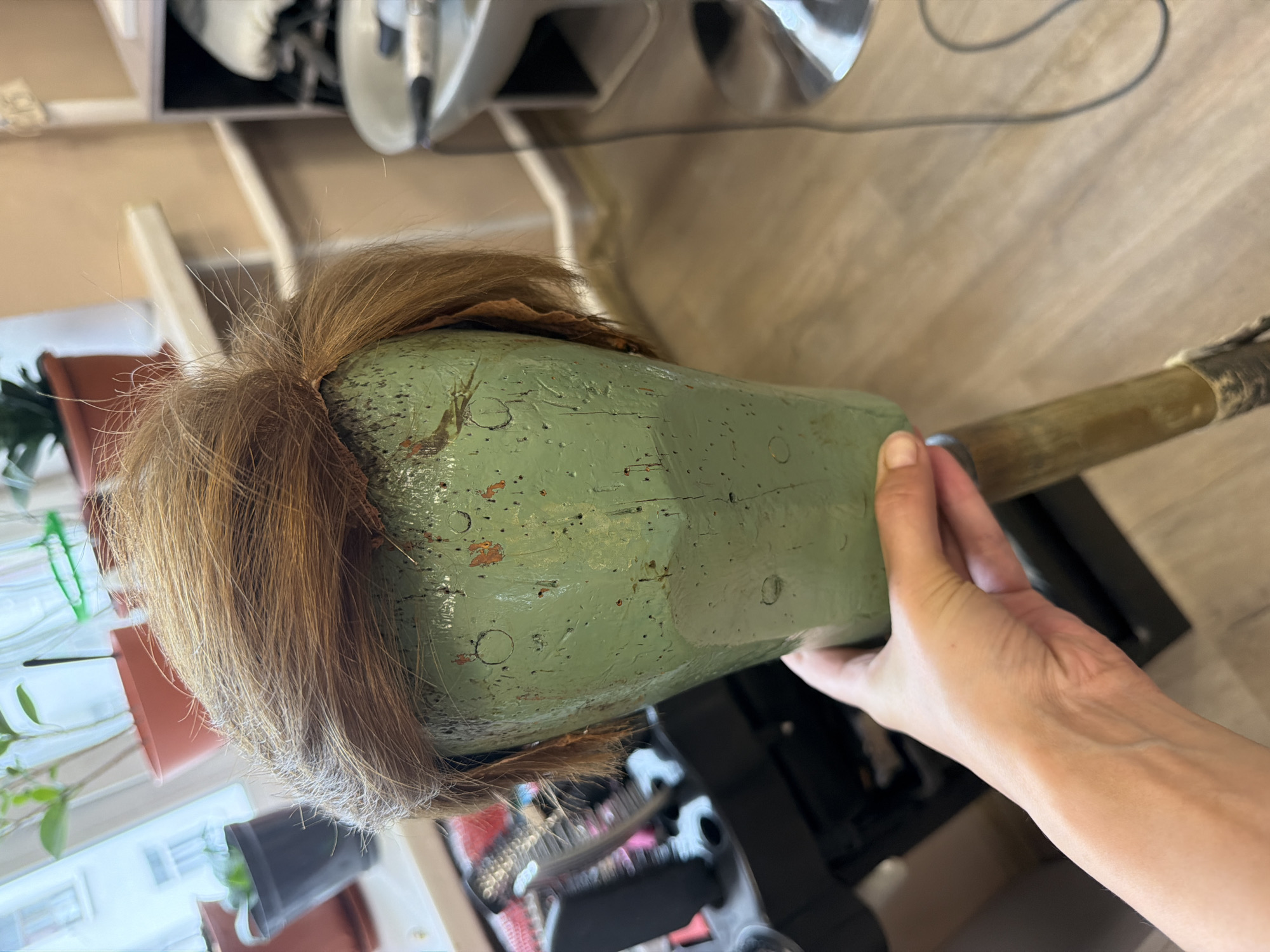 A human hand is holding a wig on a green mannequin head, which is mounted on a wooden stand. The wig is a reddish-brown, short, and appears to be styled with some flyaway hairs. The mannequin head is worn and chipped, with visible paint damage. In the background, a window with potted plants can be seen, as well as salon equipment including a chair and a stand with brushes. The flooring is light-colored wood. The scene suggests a hair salon or a wig styling setting. The image does not provide any specific information about the city of Khabarovsk, Russia.