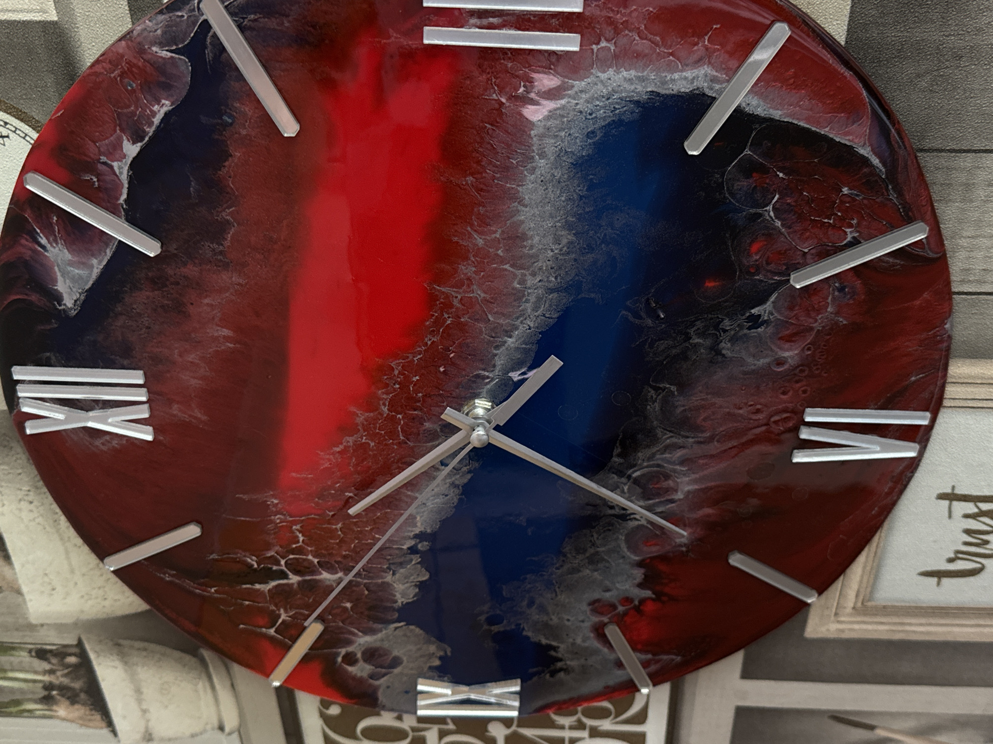 The image shows a close-up of a round wall clock with a red, white, and blue abstract resin design on its face. Silver-colored, rectangular markers indicate the hours. Roman numerals are used for 11 (XI) and 5 (V). The clock's hands are silver, with the minute hand pointing to approximately 4:20. The clock appears to be wall-mounted. A framed picture, with the word "trust" visible, is partially visible in the lower right corner. Other blurred decorative elements can be seen in the background. The scene does not appear to be in Izhevsk, Russia, as there is no identifiable context related to the location.