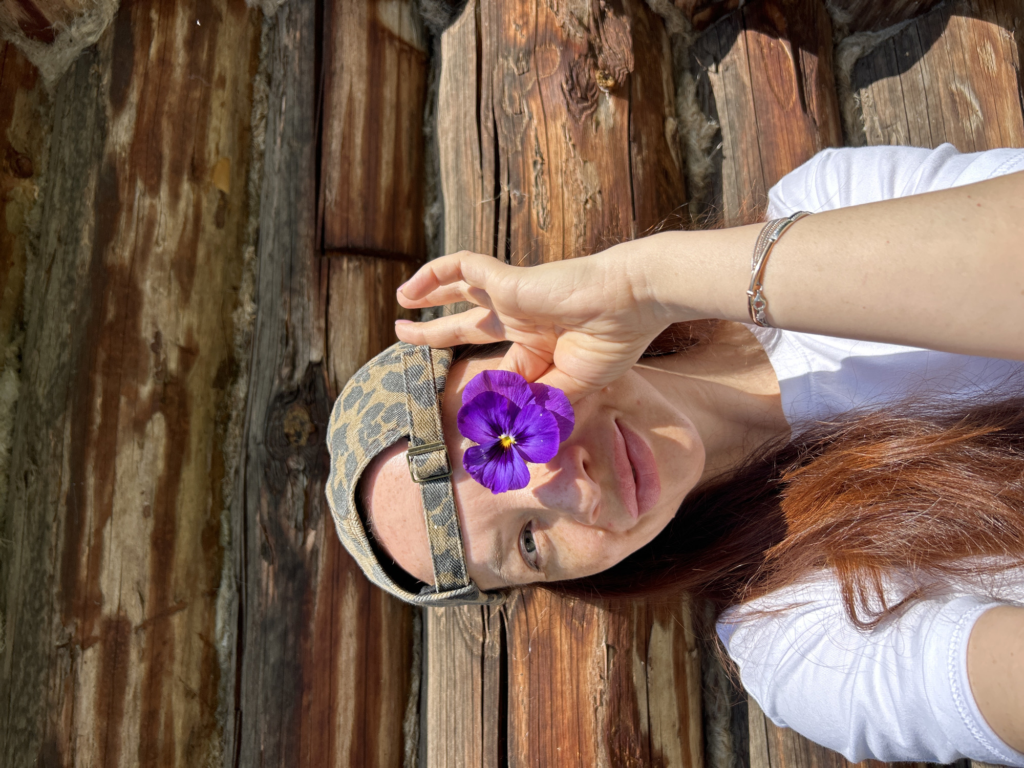A close-up, slightly low-angle, outdoor, waist-up selfie of a woman with auburn hair and fair skin, freckles, and light green eyes. She is wearing a white t-shirt, a leopard-print baseball cap, and a silver bracelet with a heart design on her left wrist. A single purple pansy flower is held in front of her right eye, obscuring it from view. Her mouth is slightly open as she smiles. She is positioned in front of a rustic, weathered log wall. The lighting suggests it is daytime, likely sunny, with shadows cast on the wall. The specific location in Chaikovsky, Russia, cannot be determined from the visual information provided in the image.