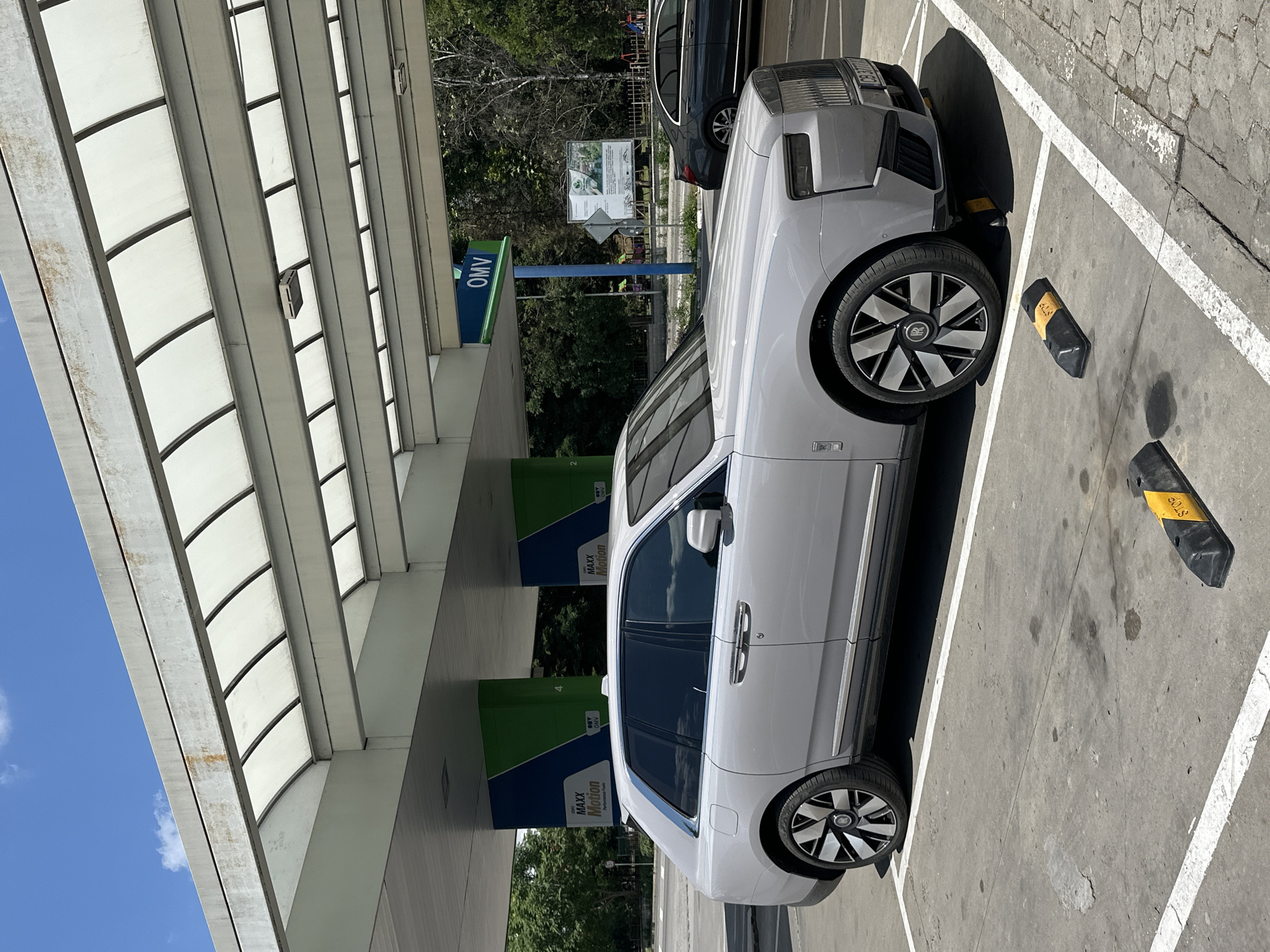 The image shows a light gray Rolls-Royce parked at an OMV gas station in Veliko Tarnovo, Bulgaria. The car is angled, with its front half facing away from the camera. The gas station canopy, made of white translucent panels supported by metal beams, covers the parking spot. Two other vehicles are partially visible: a dark-colored sedan behind the Rolls-Royce, and a glimpse of another car beneath the canopy.
In the background, there are trees and a blue sky with a few white clouds. A sign with the OMV logo and "MAXX Motion" is visible on one of the gas pumps, which are colored green and blue. Another sign behind the Rolls-Royce has text and images, suggesting information about the area or services.
The ground is concrete, marked with white parking lines and a few dark stains. Two black wheel stops, one with a yellow label that reads "STOP," are positioned near the Rolls-Royce's tires. The sunlight suggests it is daytime, likely mid-morning or afternoon, given the shadows. The scene appears to be a typical gas station stop. No people are clearly visible in the frame.