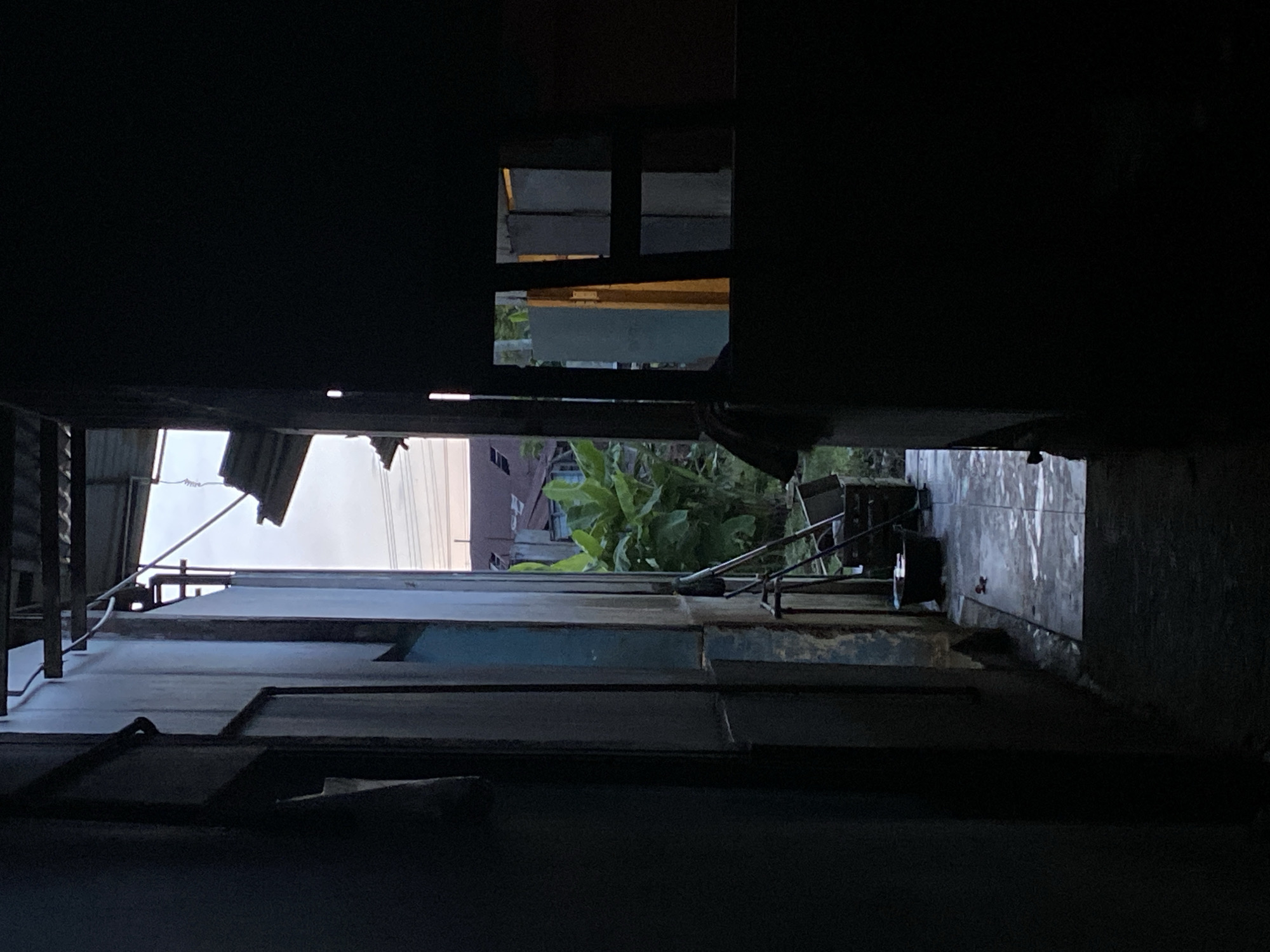 The image depicts a narrow, dark alleyway in Gampongaceh, Indonesia. The view is looking down the alley, framed by the eaves and walls of adjacent buildings. The buildings appear to be constructed of corrugated metal and weathered wood. There are visible openings or windows in the structures, allowing glimpses of greenery and a building further down the alley. The floor of the alleyway is tiled and damp. Debris is seen in the middle ground. The overall impression is one of a cramped, shadowy space with a sense of decay.