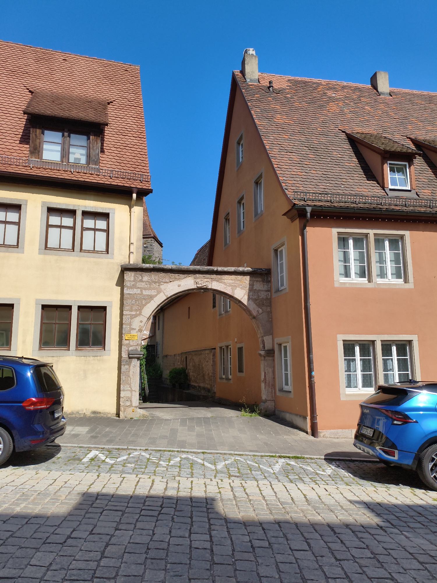 The image depicts a street scene in Rothenburg ob der Tauber, Germany, specifically in the vicinity of the Plönlein, a famous triangular intersection. Two buildings flank a stone archway that leads to a narrow street lined with more buildings. The building on the left is painted yellow with traditional wooden-framed windows. The building on the right is painted a warm orange hue, also featuring multiple windows. A blue SUV is parked on the cobblestone street in the foreground, partially obscuring the view of the archway's entrance. The sky above is clear and blue, indicating a sunny day. The architecture suggests a historical European town setting.