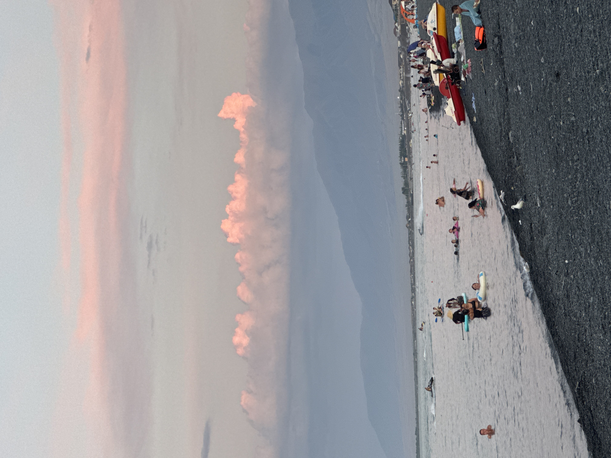 The image depicts a scenic view of a beach on a dusky day, likely at the resort village of Ақши, Kazakhstan, situated on the shores of Lake Balkhash. The sky is painted with soft pink and pale blue hues, suggesting the sun is either rising or setting, with fluffy clouds adding texture to the upper portion of the frame. In the foreground, a dark, pebbly beach is visible, with several people scattered across it. Some are seated, while others are standing, and a few are lying down. A white bird is present on the beach. Adjacent to the beach, several colorful pedal boats, red and yellow, are parked. The water of Lake Balkhash extends into the midground, populated with numerous individuals engaged in various water activities. Some are swimming or wading, while others are on paddleboards or small watercraft, including what appears to be a jet ski in the distance. Further out, a line of people are visible in the water, some swimming and others on inflatable rings. In the background, the shoreline is lined with buildings and vegetation, indicating a populated area. Beyond the shoreline, a range of hazy, blue mountains forms the backdrop, with the sky meeting the peaks in a soft gradient. The overall atmosphere is one of leisure and recreation on a tranquil evening.