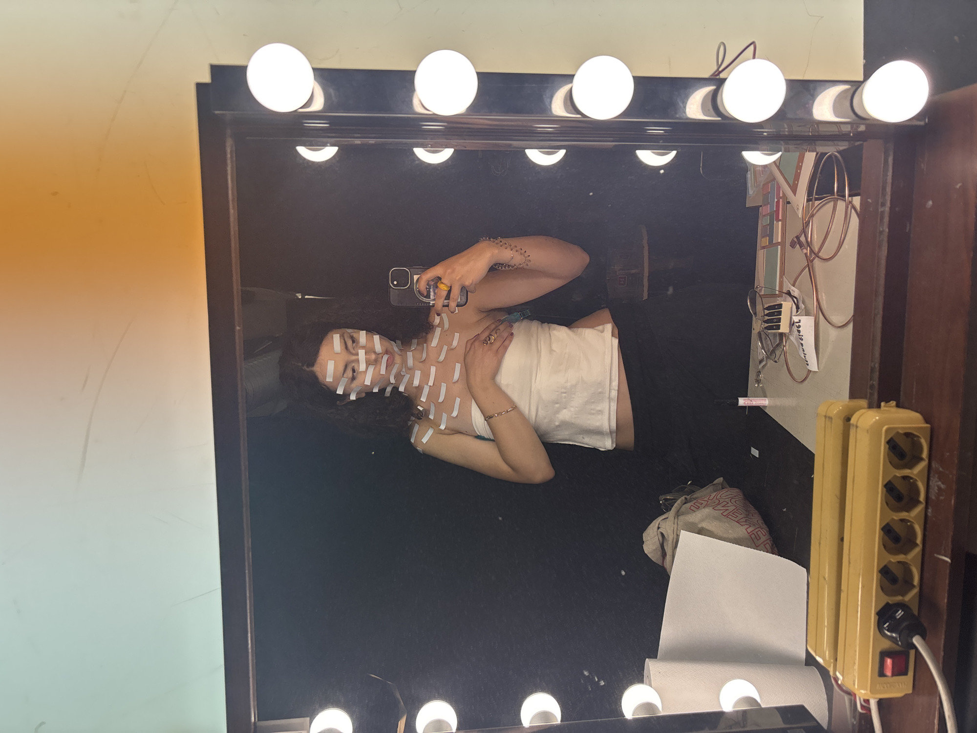 A young woman is lying down in front of a mirror, taking a selfie. She has long, dark curly hair and is wearing a white strapless top and black pants. Her body is covered in numerous white pieces of tape. She is wearing rings on her fingers and a bracelet on her wrist. Her expression is neutral. The setting appears to be a dressing room or makeup area, with a large mirror surrounded by lights. Various objects are visible in the reflection of the mirror, including a yellow power strip with multiple outlets and a red switch, some cables, a roll of paper towels, and a bag. Behind the woman, the reflection shows a dark surface. The lighting is bright due to the numerous bulbs around the mirror, suggesting it's likely daytime or an indoor setting with artificial lighting. The location is identified as Milan, Italy. There is no discernible weather information. Some faint lines and smudges are visible on the mirror itself.