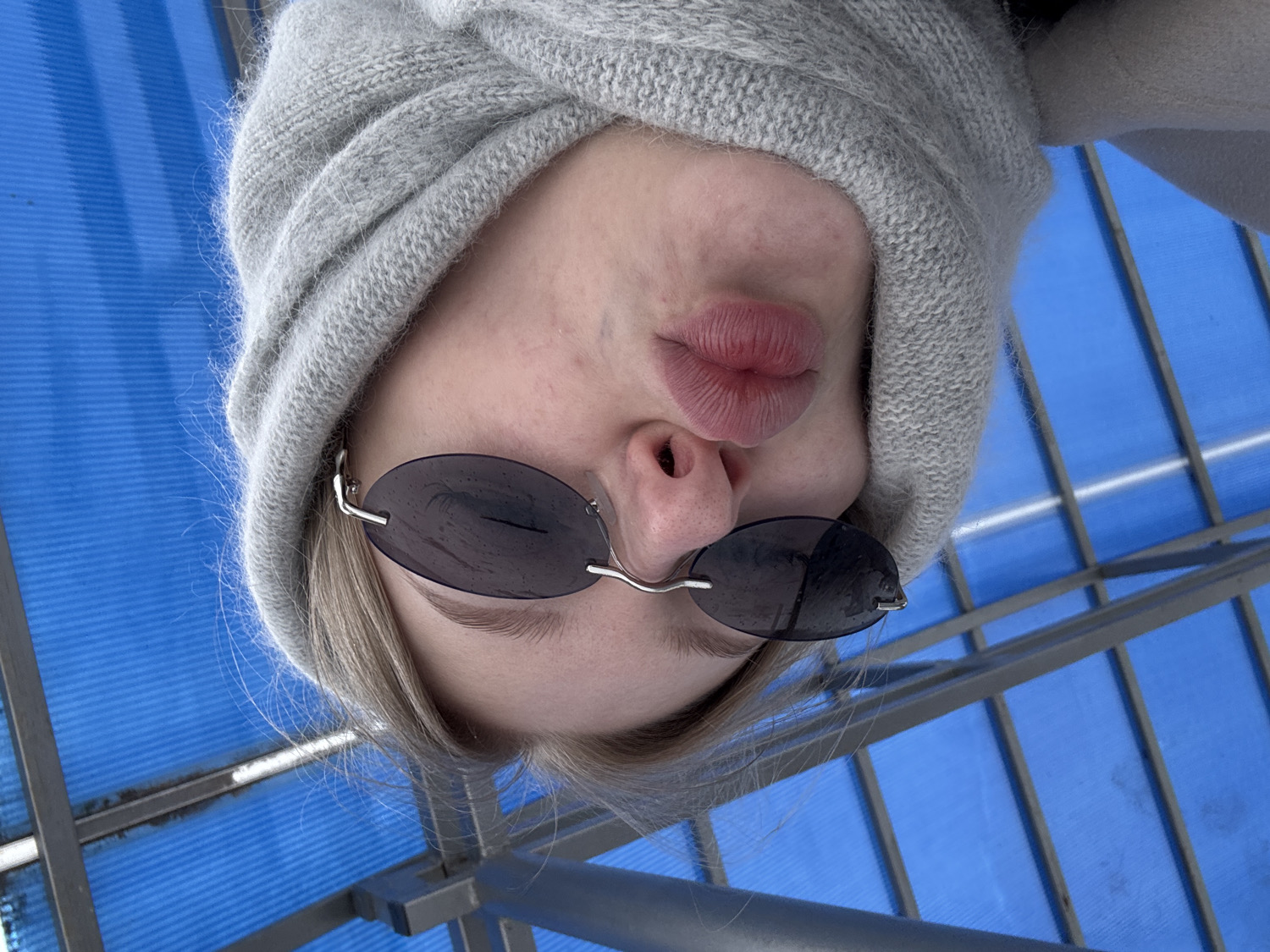 A close-up, low-angle photograph captures a person's face looking directly down at the camera, appearing upside down against the background. The individual, likely female, wears a soft, light grey knitted beanie or hat that frames their face, with light-colored hair visible underneath. They have small, oval-shaped dark sunglasses with thin silver frames resting on their nose. The person's lips are visibly pursed in a playful "kiss" expression, and the lower part of their face, from the nose to the chin, is in focus. Some minor skin texture and small blemishes are visible on the skin around the nose and mouth.
The background features a distinct, bright blue structure, possibly a translucent ceiling or canopy, supported by a grid of grey metallic beams, creating a geometric pattern behind the subject. The lighting appears bright and even, suggesting a daytime setting. The overall impression is one of a casual and expressive moment. No text is visible in the image. The location context for this image is Perm, Russia.