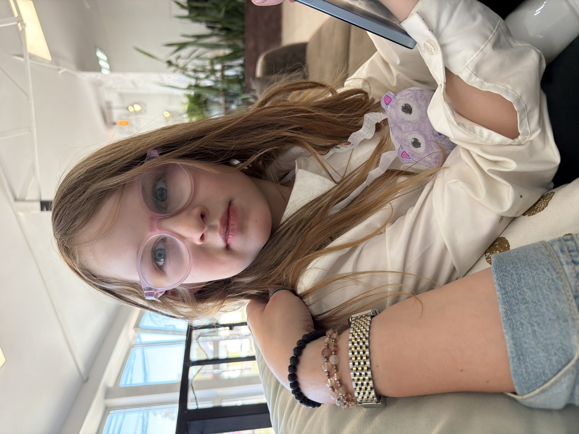 A young girl with light brown hair and pink-rimmed glasses is looking at the camera. She is wearing a white shirt and has a drawing of a purple bear pinned to her chest. She is holding a black tablet. The girl is sitting on a light-colored patterned sofa. Her arm is visible, adorned with a watch and two beaded bracelets. In the background, there's a window with light streaming through, a potted plant, and a glimpse of a modern interior. The location is Chaikovsky, Russia.