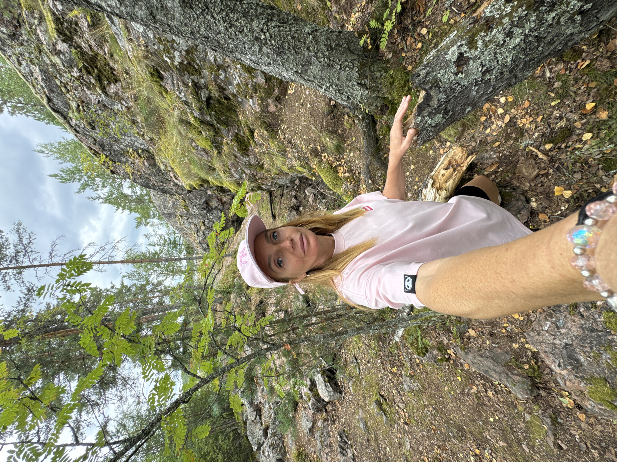 A woman in a pink baseball cap and pink t-shirt is taking a selfie in a wooded, rocky area. She has blonde hair and is wearing a bracelet with pink and clear beads. Her left arm is visible in the foreground, holding the camera. The background shows a rocky incline covered in moss and grass, with trees and a cloudy sky above. The ground is covered in dirt, rocks, and fallen leaves. The location is identified as the city of Sorola, Russia.