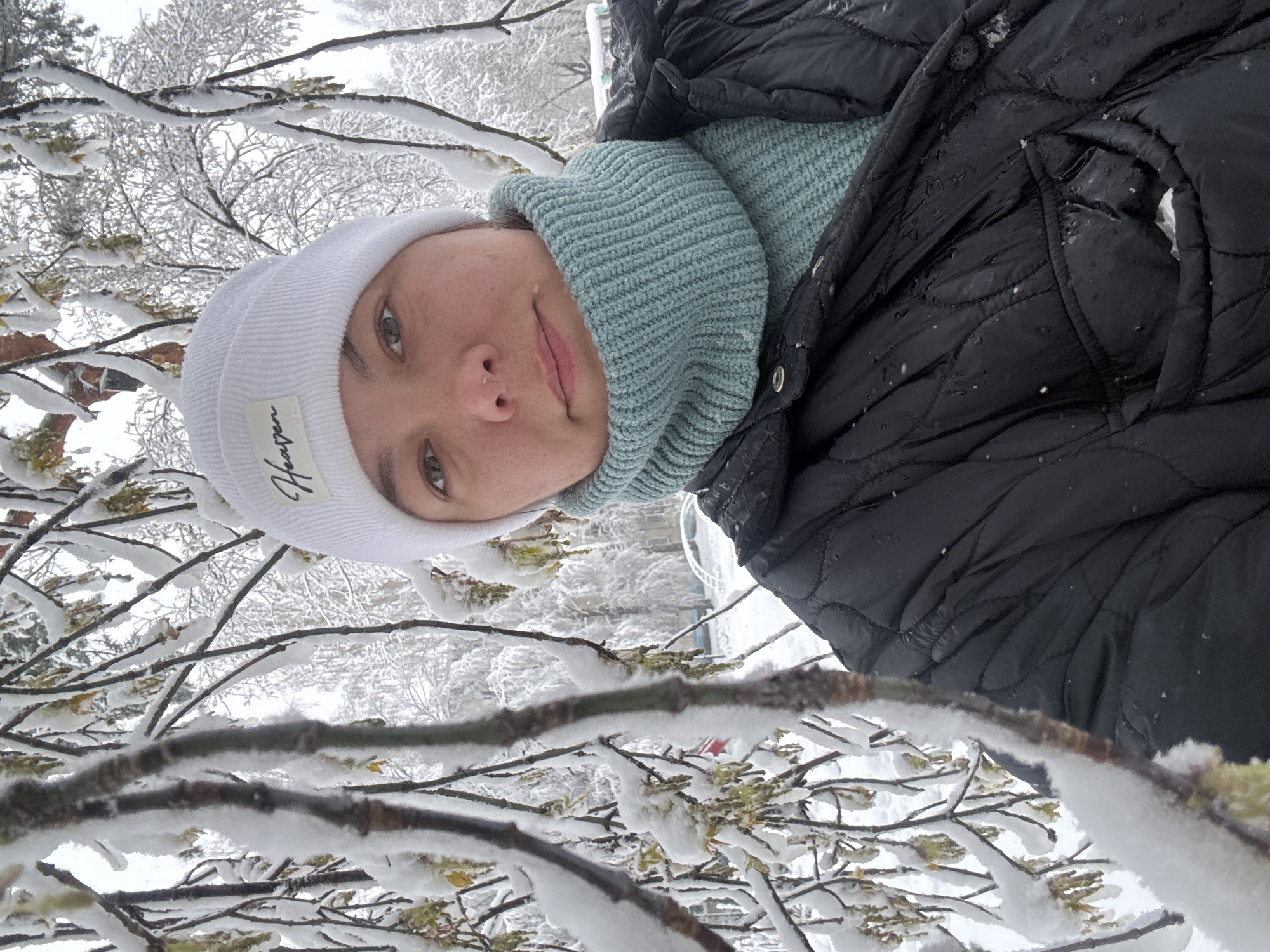 A close-up, eye-level selfie captures an individual in a snowy outdoor environment in Dmitrov, Russia. The subject, likely a young adult, is seen from the chest up, with their head slightly tilted. They wear winter clothing including a white ribbed beanie, a light blue chunky knit snood or neck warmer, and a black quilted winter jacket. A faint, gentle smile plays on their lips, and they look directly towards the camera with light-colored eyes.
The foreground and background are filled with tree branches heavily covered in fresh, white snow, creating a dense, wintry scene. Some small green or yellowish buds/leaves are visible on the snow-laden branches, indicating plant life beneath the snow. The black jacket shows small water droplets or melting ice, suggesting recent snowfall or slightly warmer temperatures causing the snow to melt. In the distance, more snow-covered trees are visible under an overcast, bright sky.
A white fabric label stitched onto the front of the beanie displays the word "Heaven" in a vertical, stylized script. The overall impression is one of a peaceful, cold, and snowy day.