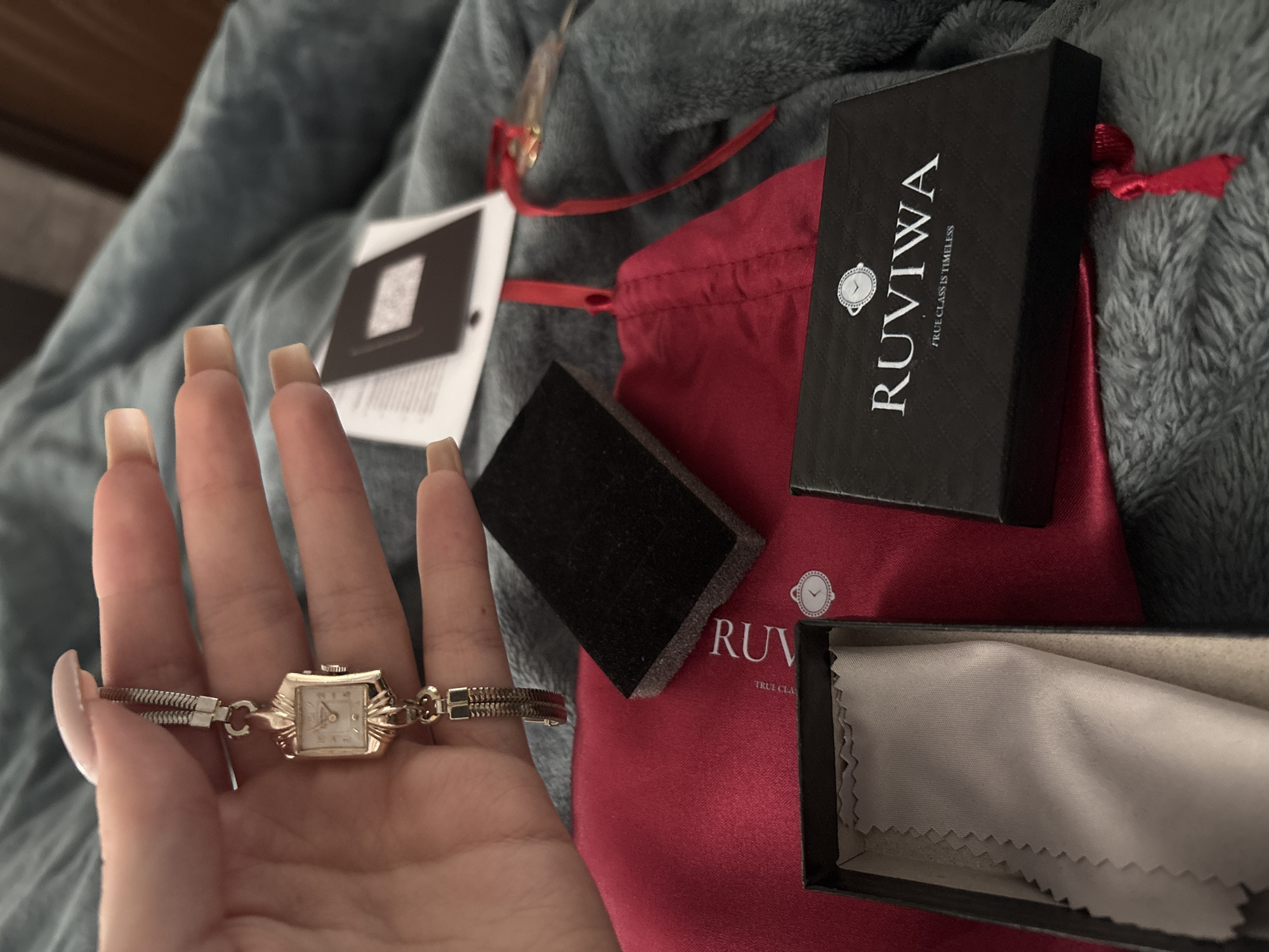 A woman's hand with long, nude-colored nails holds a gold wristwatch with a rectangular face. The watch band appears to be a braided metal design. Behind the hand, resting on a textured gray surface that appears to be a blanket or throw, are several items associated with the wristwatch: a black box labeled "RUVIWA / TRUE CLASS IS TIMELESS" atop a red drawstring pouch, a small black square of what might be foam or velvet, and an opened black box containing a light-colored cleaning cloth. A white tag attached with a red string is partially visible in the background. The image was not taken in Sofia, Bulgaria.