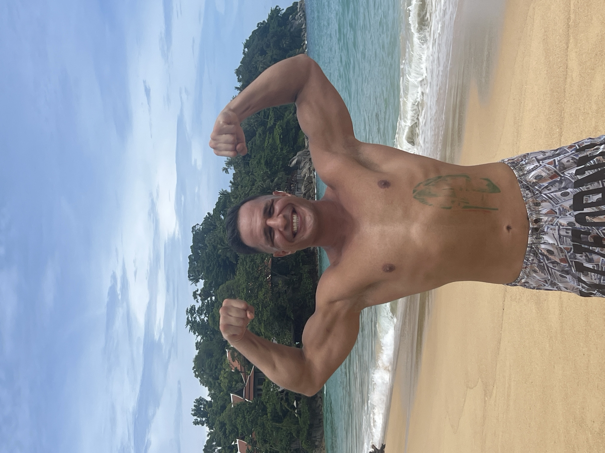 A man is standing on a beach in Karon, Thailand, flexing his biceps. He is wearing patterned shorts and has a tattoo on his left side. The ocean is behind him with waves breaking on the shore, and lush green trees and buildings are visible on the hillside in the background. The sky is cloudy.