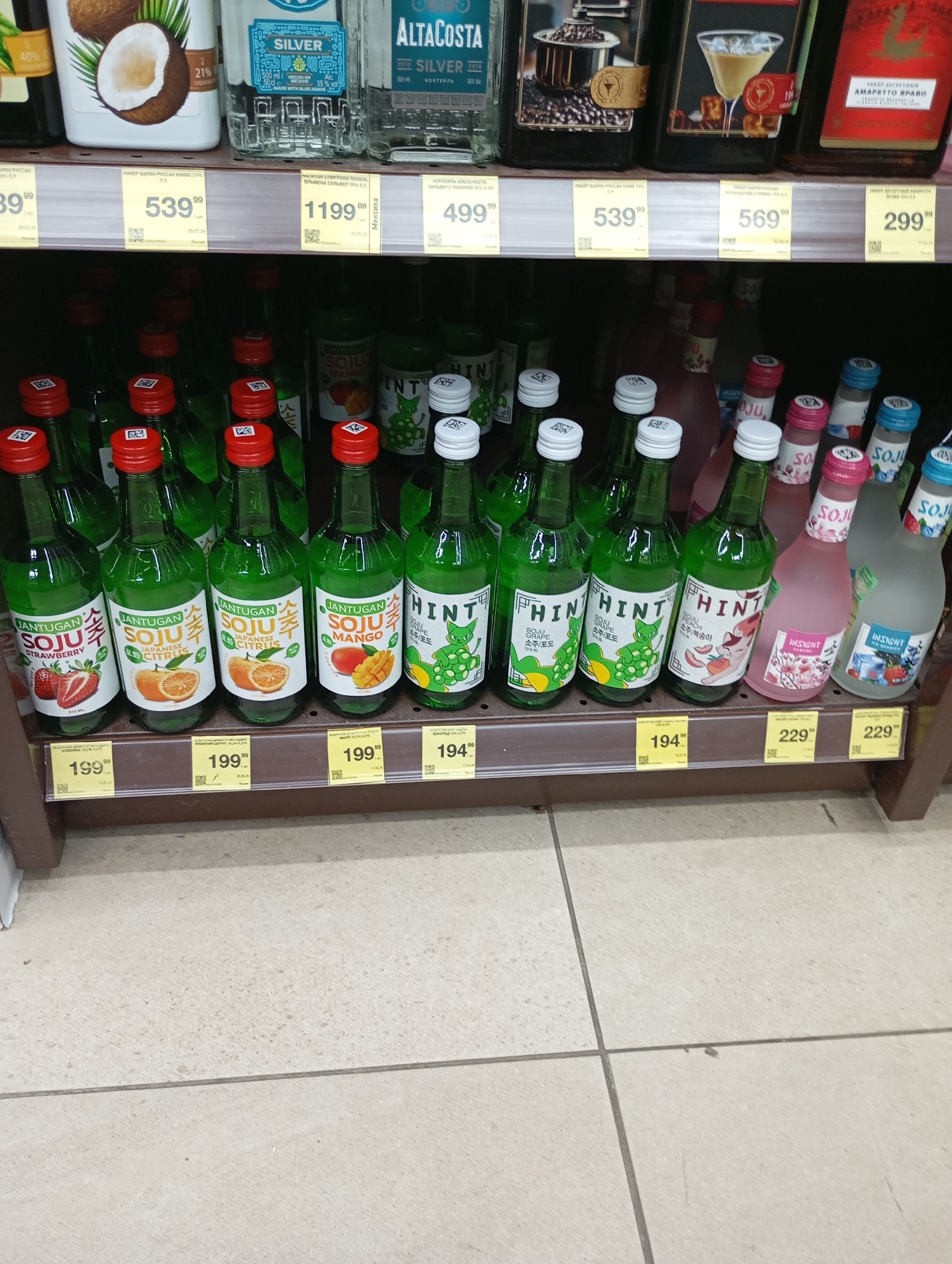 The image shows a retail shelf display in a Moscow, Russia store. The shelves are stocked with various bottles of alcoholic beverages, predominantly soju and amaretto. The bottles are arranged in rows on two levels. The lower level features small, green glass bottles of soju with colorful labels indicating flavors such as strawberry, Japanese citrus, mango, grape, and peach. Some of these bottles have red or white caps. To the right of the soju bottles are two larger, frosted glass bottles, one pink and one clear, also containing soju. The upper level displays larger bottles of liquor, including a coconut-flavored liqueur, several bottles of "Silver" branded spirits, and amaretto. Price tags are visible on the shelf edge, indicating prices in Russian Rubles. The background is a retail store environment with neutral-colored flooring and shelving units. There are no people or animals visible in the image. The lighting appears to be overhead retail lighting.