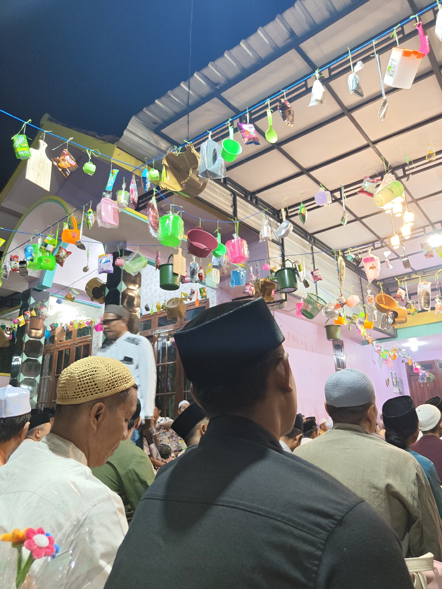 The image captures an outdoor gathering in Cermee, Indonesia, possibly for a religious event or celebration, given the presence of many men wearing peci hats and observing a speaker. The scene is decorated with numerous colorful plastic household items like cups, buckets, and small containers, suspended from strings across the roof of a structure. These items appear to be part of a festive or symbolic display. The roof structure itself is made of metal and supported by metal beams. In the foreground, the backs of several men are visible, with one man in a black shirt and peci facing away from the camera. Another man in a white shirt and a tan woven peci is seen from the side, looking down. Further back, a man in a white shirt and glasses is visible. The background reveals a building with wooden doors and windows. The lighting suggests it is evening or night, with artificial lights illuminating the area.