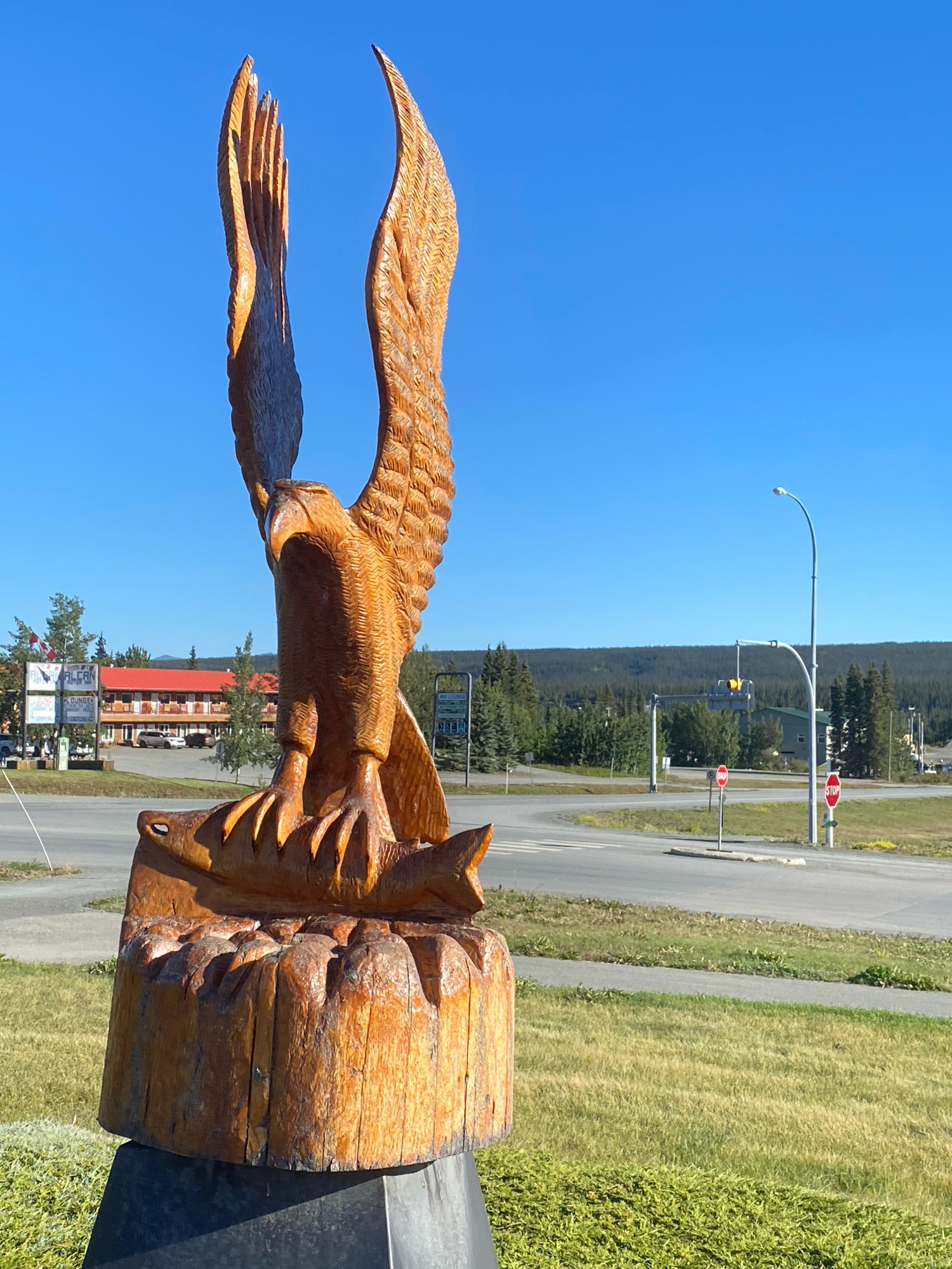 A large, intricately carved wooden sculpture of an eagle with outstretched wings, perched on a base that depicts a fish and a log, stands prominently in the foreground. The sculpture is made of light-brown wood and is positioned outdoors under a clear blue sky. In the background, a road with multiple lanes curves through a grassy area, bordered by trees and some buildings. A red-roofed hotel with the sign "ALCAN MOTEL LOUNGE" is visible to the left, along with a traffic light and stop signs on the right. The overall setting appears to be a roadside or town square in Haines Junction, Canada, likely a tourist area given the prominent display of the sculpture. The environment is sunny and calm, with no people visible in the frame.