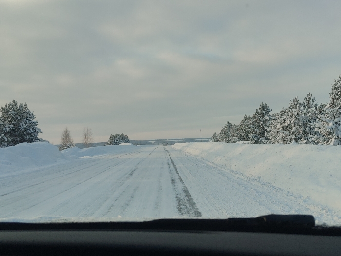 A snowy road stretches ahead under a gray, overcast sky. Both sides are lined with snow-covered trees and deep banks of snow. Tire tracks are visible on the road surface, indicating recent travel. The view is from inside a vehicle, with part of the dashboard visible in the lower portion of the frame. The scene evokes a sense of quiet, isolated travel through a wintry landscape.