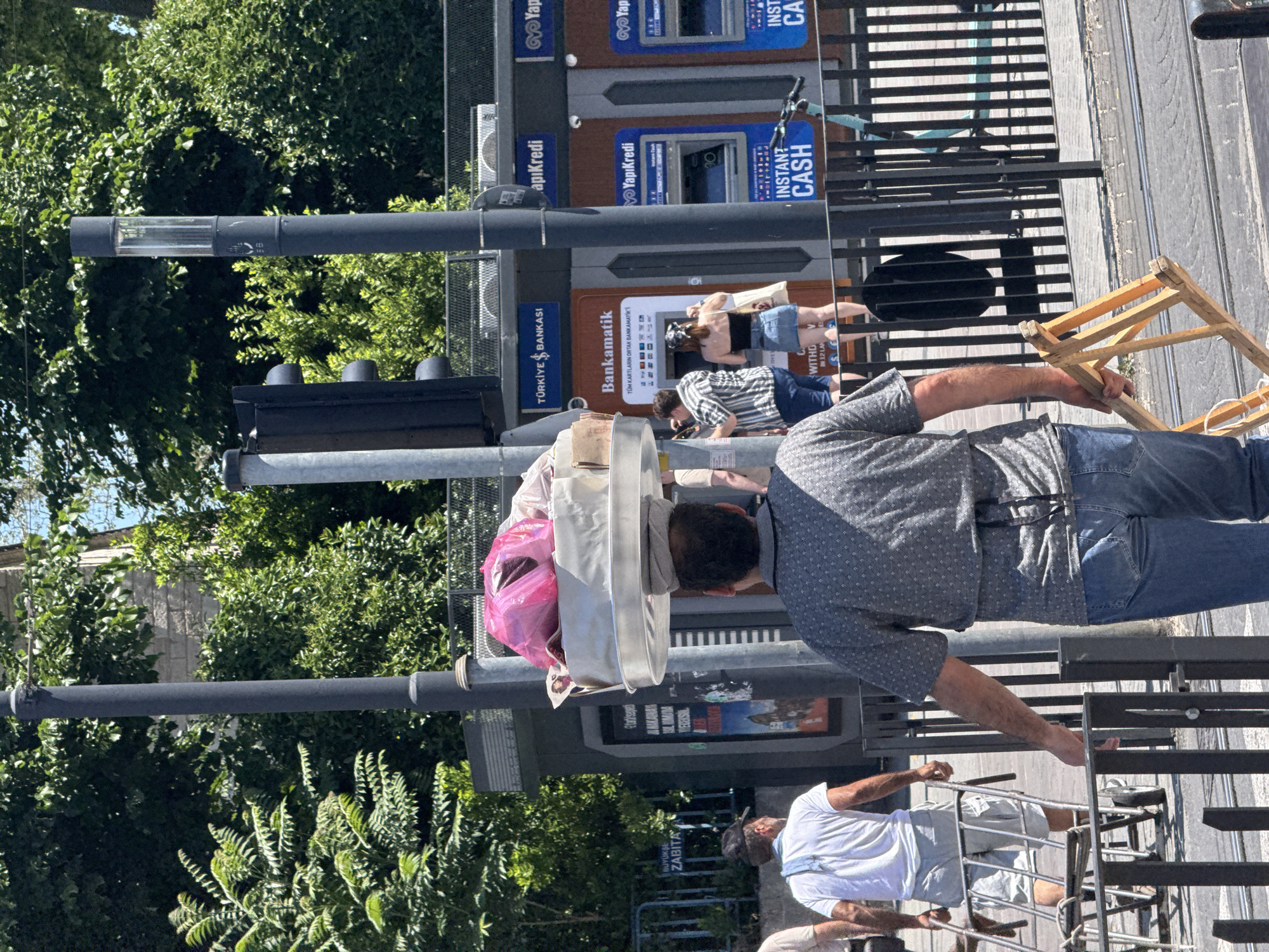 The image depicts a sunlit urban street scene in Istanbul, Turkey. In the foreground, viewed from behind, a man with dark hair carries a stack of items, including a prominent pink plastic bag, balanced on a tray on his head, supported by a white cloth. He is wearing a grey patterned short-sleeved shirt and blue jeans, and holds a light-colored wooden frame in his right hand. To his left, another man, also viewed from behind, wears a white t-shirt, light shorts, and a dark baseball cap, pushing a metal hand truck or cart. A white cloth is draped over his left shoulder.
In the mid-ground, a row of automated teller machines (ATMs) is visible. These include "Türkiye İş Bankası" and "YapıKredi" branded units, featuring brown and blue enclosures, respectively. Two individuals, a man in a striped shirt and a woman in a black top and denim skirt with a bandana, are standing in front of the "Türkiye İş Bankası" ATM. A light blue electric scooter is parked near the "YapıKredi" ATMs, behind black metal street railings.
The environment includes a paved street with visible tram tracks in the foreground. Dark grey lampposts, one featuring a hand-like symbol and numbers "56", line the street. A black traffic light is visible near the ATMs. Green foliage, comprising numerous trees and bushes, dominates the background, partially obscuring a light-colored stone building. A dark grey sign with "BÜYÜKŞEHİR ZABITA" (Metropolitan Municipal Police) is visible on the far left. Air conditioning units are mounted above the ATM structure. The scene is bright, indicating clear weather.