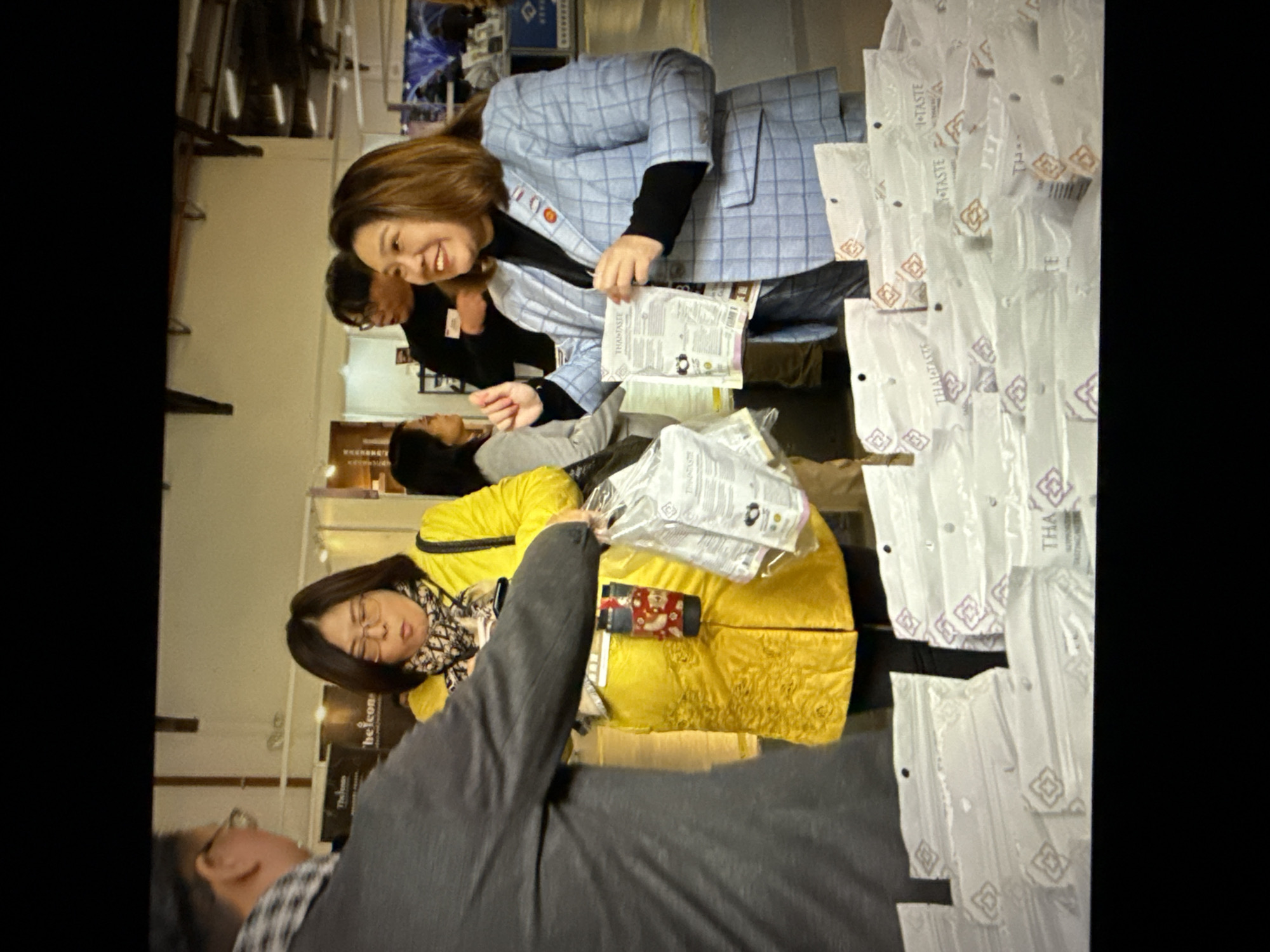 In a dimly lit indoor setting, possibly a convention or trade show, a group of people are gathered around a table displaying stacks of packaged goods. A woman in a bright yellow coat, wearing glasses and a patterned scarf, is holding a bag of the products. To her right, another woman in a light blue plaid blazer, smiling and wearing a pin on her lapel, is also holding a product bag and appears to be interacting with someone off-camera. In the foreground, a large display of the packaged goods, branded with "THA TASTE", is visible. The background shows more exhibits and people, suggesting a busy commercial event. The overall atmosphere appears to be one of product promotion or sales.