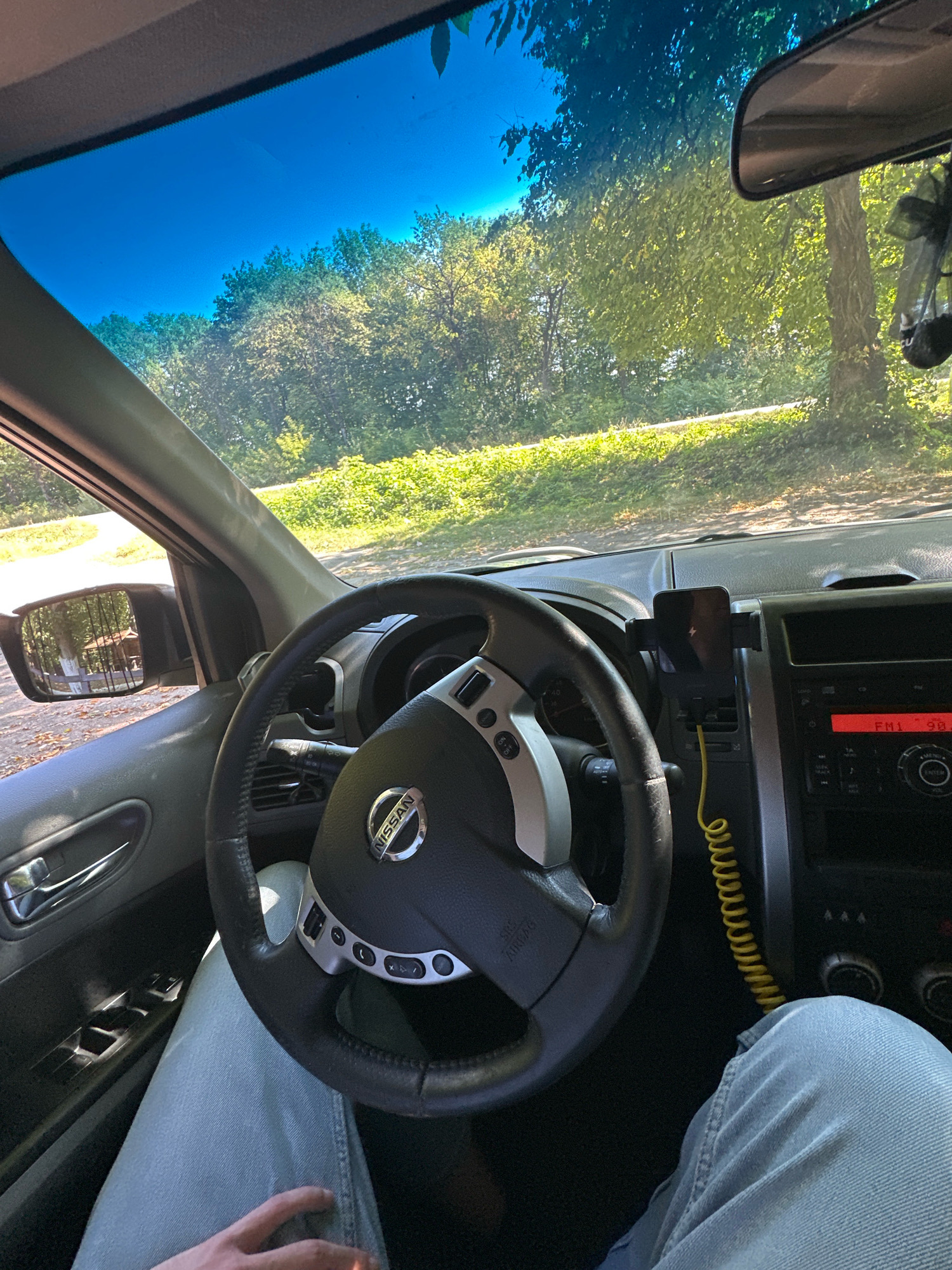 The image captures the interior of a Nissan car, with the steering wheel and dashboard in the foreground. The driver's legs, clad in light blue jeans, are visible in the lower part of the frame. A cell phone is mounted on a holder on the dashboard. Through the windshield and side window, a sunlit, tree-lined outdoor environment is visible, suggesting a rural or park-like setting. The rear-view mirror shows a reflection of trees and a small building, possibly a gazebo. The scene is illuminated by natural daylight. The location is identified as Shatava, Ukraine.