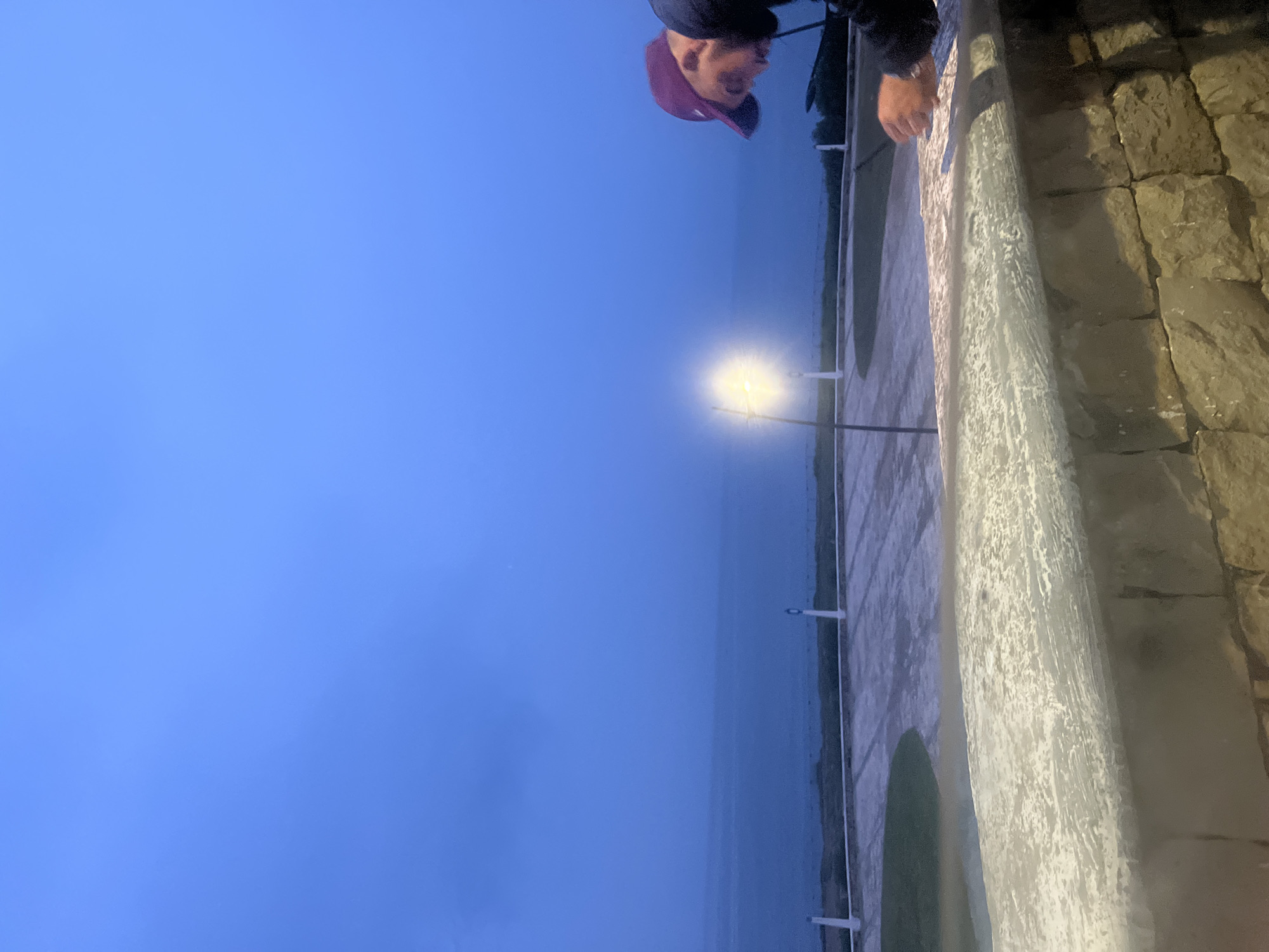 The photo shows a person wearing a red baseball cap and a dark jacket leaning over a stone wall in Fnideq, Morocco. The wall appears to be part of a coastal promenade or overlook. Beyond the wall is a body of water (likely the sea), and the sky is a hazy blue, suggesting twilight or overcast conditions. A lamppost with a bright light is visible near the water's edge. The wall is constructed from irregularly shaped stone blocks. The camera angle suggests the photo was taken from a low vantage point.