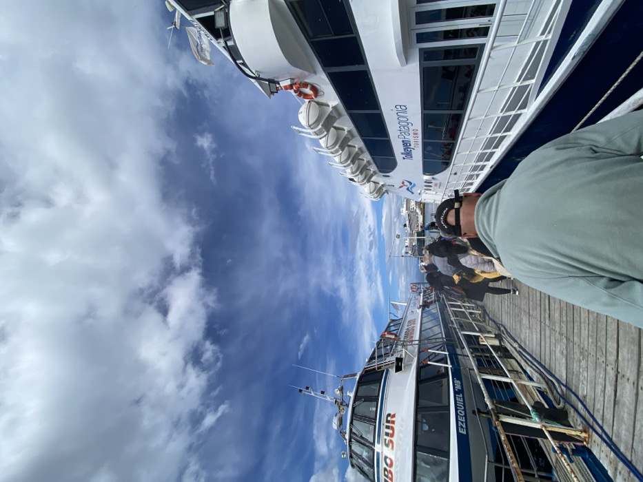 The image captures a vibrant scene at a harbor in Ushuaia, Argentina, likely during daytime under a partially cloudy, blue sky. The photo is taken from a low angle, looking upwards and across a wooden pier where several people are gathered.
In the foreground, the back of a person wearing a light green long-sleeved top and a black baseball cap is visible, facing towards the water. Beyond this person, a group of other individuals, mostly seen from behind, are walking along the pier, suggesting an activity involving embarkation or disembarkation from boats.
Two large passenger boats, or catamarans, are prominently docked side-by-side. The boat closer to the camera is white and blue, with the name "RUMBO SUR" clearly visible on its side, along with "EZEQUIEL 'MB'". It features multiple large windows and antennas on its roof. The second boat, positioned further back and slightly higher in the frame, is also white and blue. On its side, the text "TOLKEYEN PATAGONIA TURISMO" is visible, indicating its purpose as a tourist vessel for the Patagonian region. This boat also has numerous windows, safety rafts stowed on its side, and an orange lifebuoy.
The bright sunlight and scattered white clouds suggest pleasant weather, ideal for boat excursions. The overall impression is that of a bustling tourist port, with people preparing for or returning from a scenic tour in the Patagonian waters.