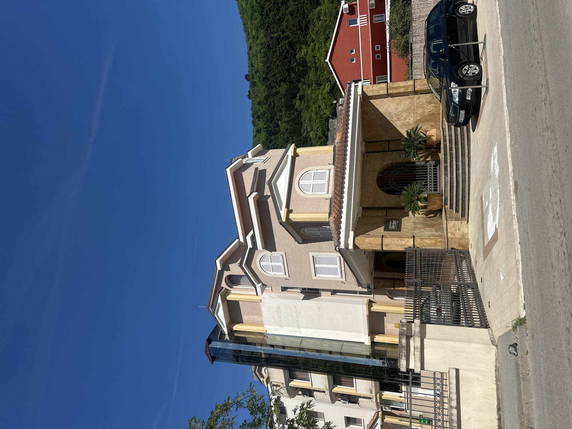 The image depicts a multi-story building in Budva, Montenegro, characterized by a beige facade with white window shutters and a distinct architectural design that includes rounded windows and decorative elements. An external glass elevator is attached to the side of the building. A black car is parked in front of the building's entrance, which features steps leading to an arched doorway adorned with potted plants. A red building is situated adjacent to the main structure, while a lush green hillside forms the backdrop. The clear blue sky suggests a sunny day. A metal fence surrounds the property, and a paved sidewalk extends along the front.