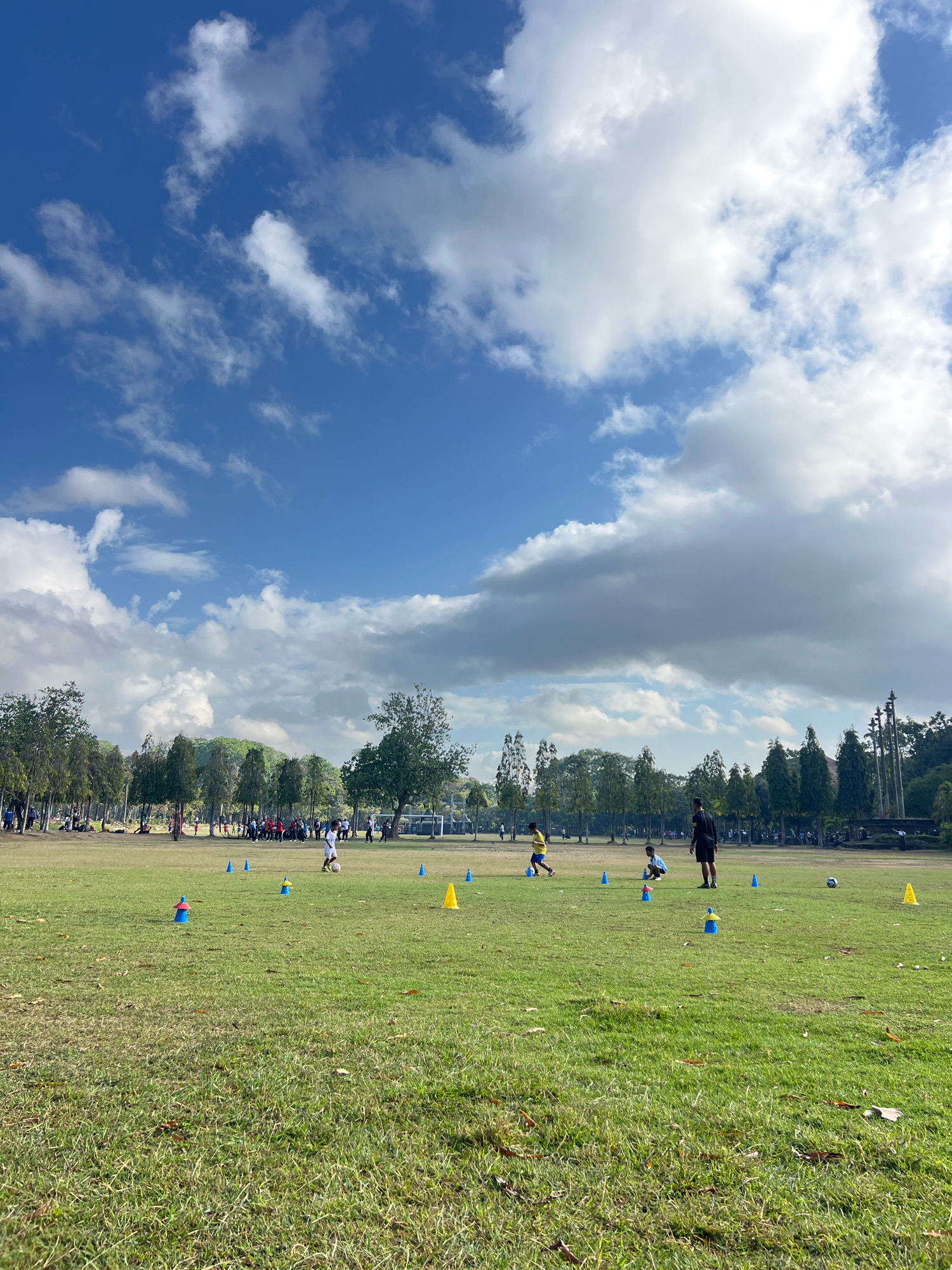 This image depicts a sunny day at a park in Denpasar, Indonesia, likely on a soccer field. The sky is a bright blue, filled with fluffy white clouds. In the foreground, a vast expanse of green grass covers the field, with scattered leaves and some dried patches. A few children are scattered across the field, some with a soccer ball, and various blue and yellow cones are set up, indicating a training session or practice. In the midground, a group of people are gathered, possibly parents or spectators, and a line of trees forms a backdrop. Further in the distance, more trees and what appear to be buildings or structures are visible. The overall atmosphere is one of outdoor activity and recreation.