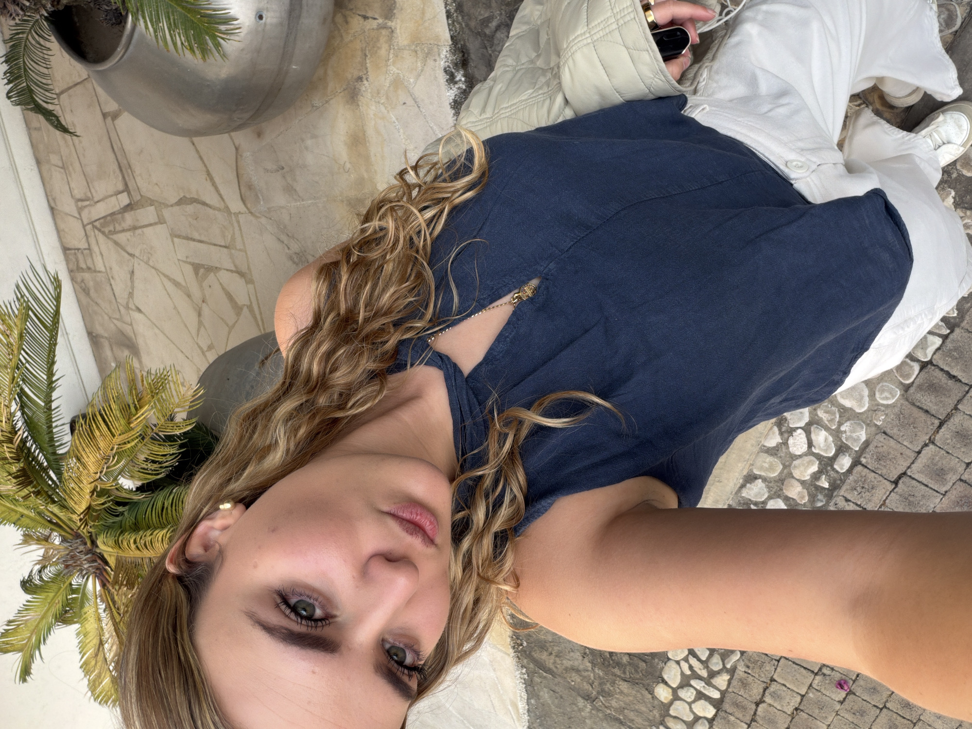 A young adult female is depicted in a high-angle selfie, looking directly into the camera. She has light skin, green eyes, and wavy light brown hair. She is wearing a dark blue sleeveless top with a keyhole neckline, which reveals a small gold-colored pendant on a chain. She wears white wide-leg pants and white sneakers. A light beige quilted jacket is draped over her left arm, and her right hand, holding a black key fob and displaying gold rings, is partially visible. The scene is outdoors in San Bernardino Tlaxcalancingo, Mexico. The ground beneath her features a mosaic of irregular light-colored flagstones and a section of cobblestone-like paving. In the background, two large metallic silver planters hold Sago Palm plants, with a light-colored wall visible behind them.