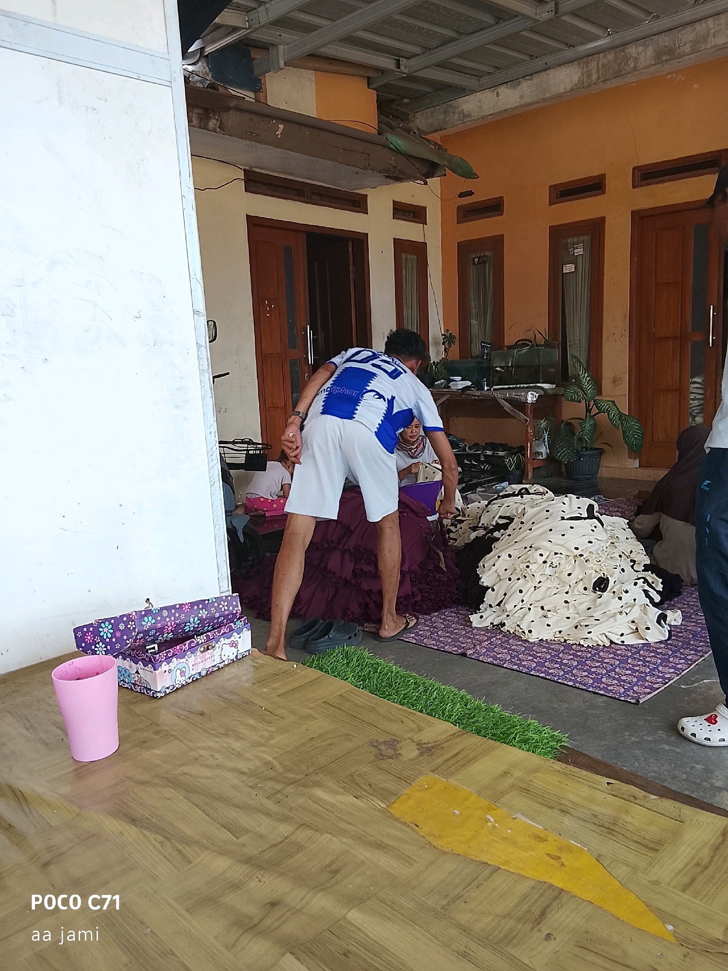 The image depicts an outdoor scene in Tasikmalaya, Indonesia, likely a residential area. In the foreground, a wooden floor with a yellow painted section is visible. To the left, a pink plastic cup and a decorative box with Hello Kitty imagery are placed on the floor.
The central figures are two individuals. One person, wearing a white and blue striped shirt and white shorts, is bent over, facing away from the camera, towards a pile of dark purple fabric. Another person, wearing a headscarf and a pink shirt, is partially visible behind the first person, appearing to be seated and looking at the fabric. A large pile of light-colored fabric with black polka dots is spread out on a purple mat to the right of the dark purple fabric.
In the background, a building with an orange-toned wall and wooden doors and windows is visible. A table with various items, including plants and what appears to be a fish tank, is situated against the building. The roof structure of the building is also apparent. The ground is paved with a grey surface in the background. A strip of artificial green grass is present between the wooden floor and the grey paved area.
The overall impression is a casual, domestic setting where laundry or fabric sorting might be taking place. The presence of the text "POCO C71" and "aa jami" in the lower left corner suggests the image was taken with a Poco C71 phone by someone named "aa jami."
