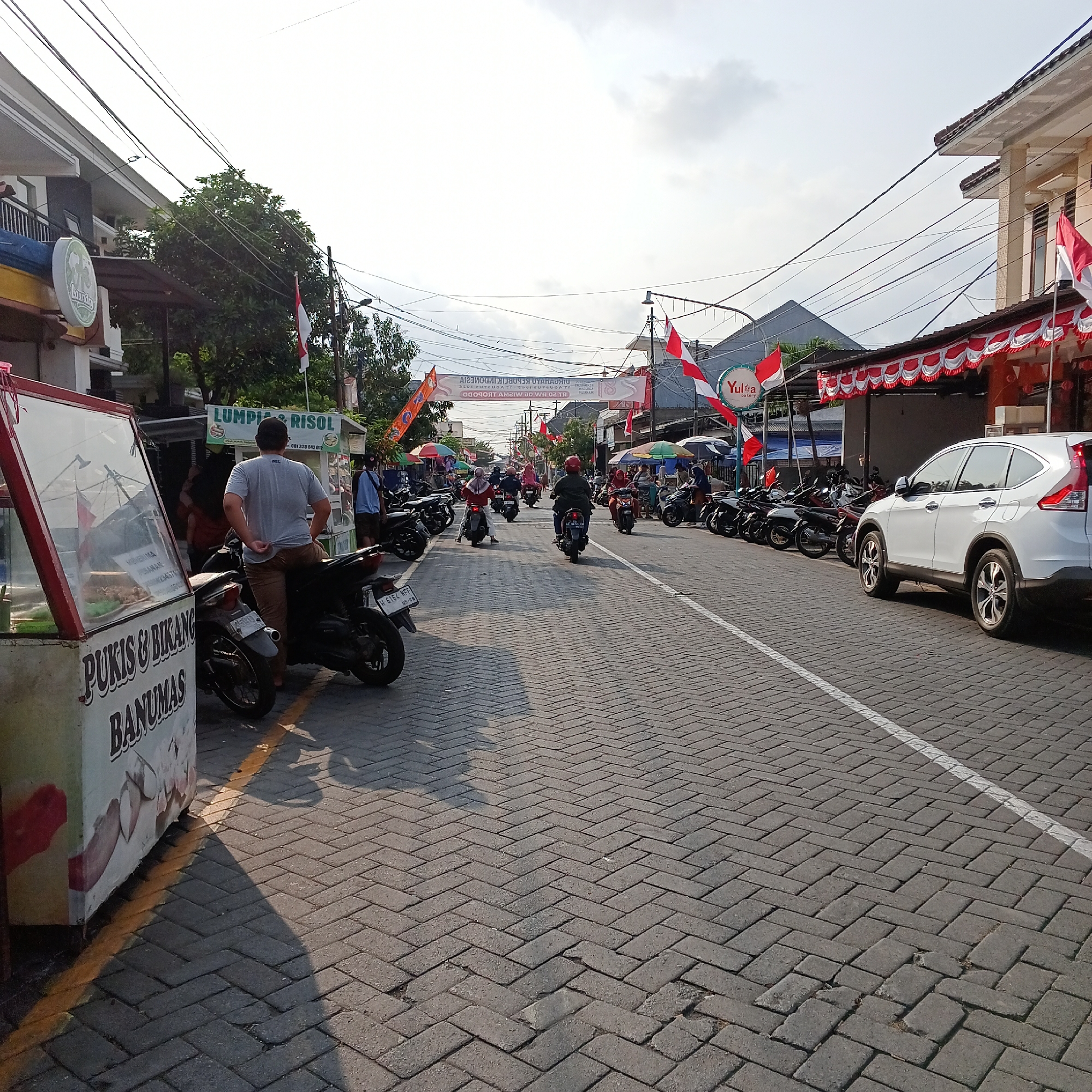 The image depicts a street scene in Tropodo, Indonesia, on a sunny day with some cloud cover. The street is paved with dark gray interlocking bricks and has a white line marking the edge of a parking area on the right. The street is lined with buildings and several parked motorcycles. On the left side, there is a food stall with a sign that reads "PUKIS & IKANG BANUMAS." A man is standing near the stall, leaning on a motorcycle. Further down the street, more motorcycles are parked, and people are seen riding them.
Several Indonesian flags are displayed along the street, along with red and white decorative banners, suggesting a local celebration or national holiday. There are also several umbrellas providing shade over stalls or seating areas. On the right side of the street, a white SUV is parked. Overhead, numerous electrical wires crisscross the sky. The overall impression is a lively street with commercial activity and local festivities.