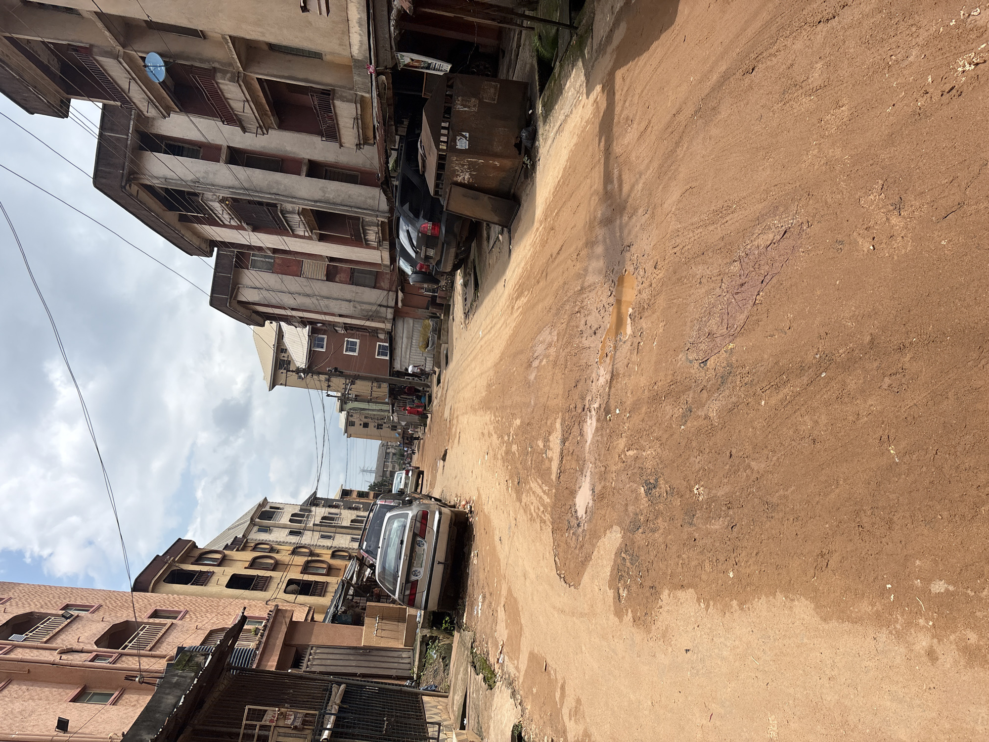 The image depicts a street scene in Onitsha, Nigeria, characterized by a red dirt road with significant potholes and puddles. Flanking the road are multi-story buildings, appearing aged and in varying states of repair, with balconies and satellite dishes visible. Several vehicles, including cars and SUVs, are parked along the street. The atmosphere is overcast, with a cloudy sky overhead. The general impression is one of an urban environment with infrastructure in need of maintenance.