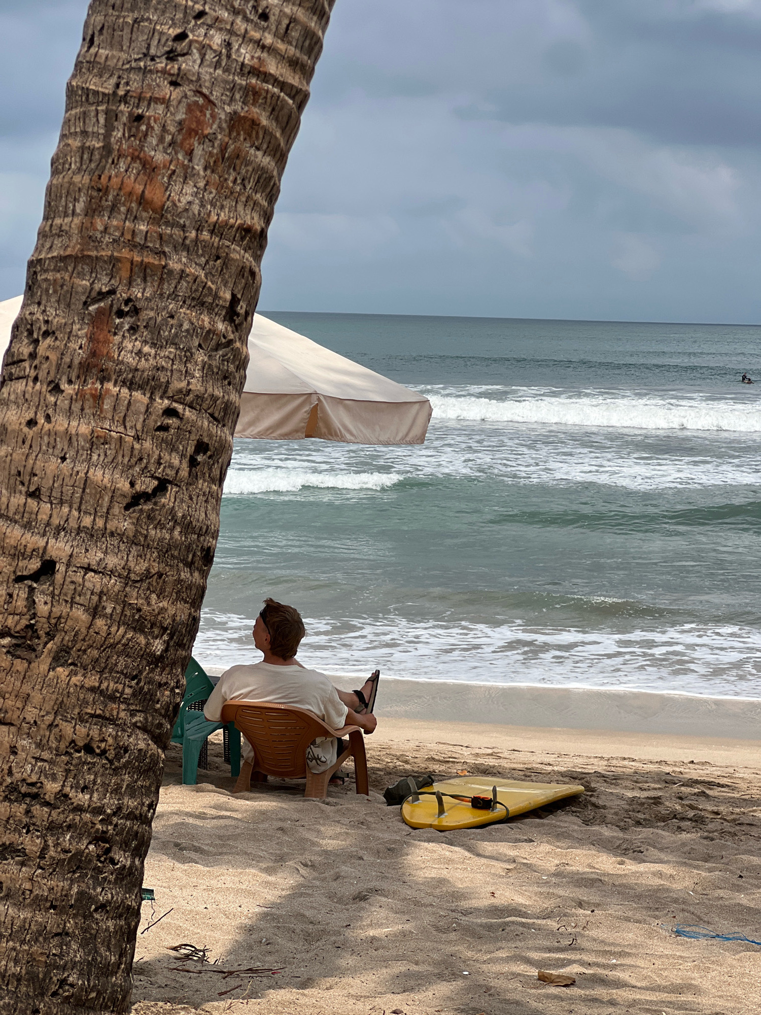 A man sits in a chair on a sandy beach in Kuta, Indonesia, facing the ocean. The man is wearing a light-colored t-shirt and shorts. To his left, a large palm tree trunk is prominently featured in the foreground, partially obscuring the view. Behind the man, a tan umbrella provides shade. A yellow surfboard lies on the sand to the right of the man. The ocean is calm with gentle waves breaking on the shore. The sky is overcast with gray clouds. In the distance, a surfer can be seen in the water. The scene appears peaceful and relaxed.