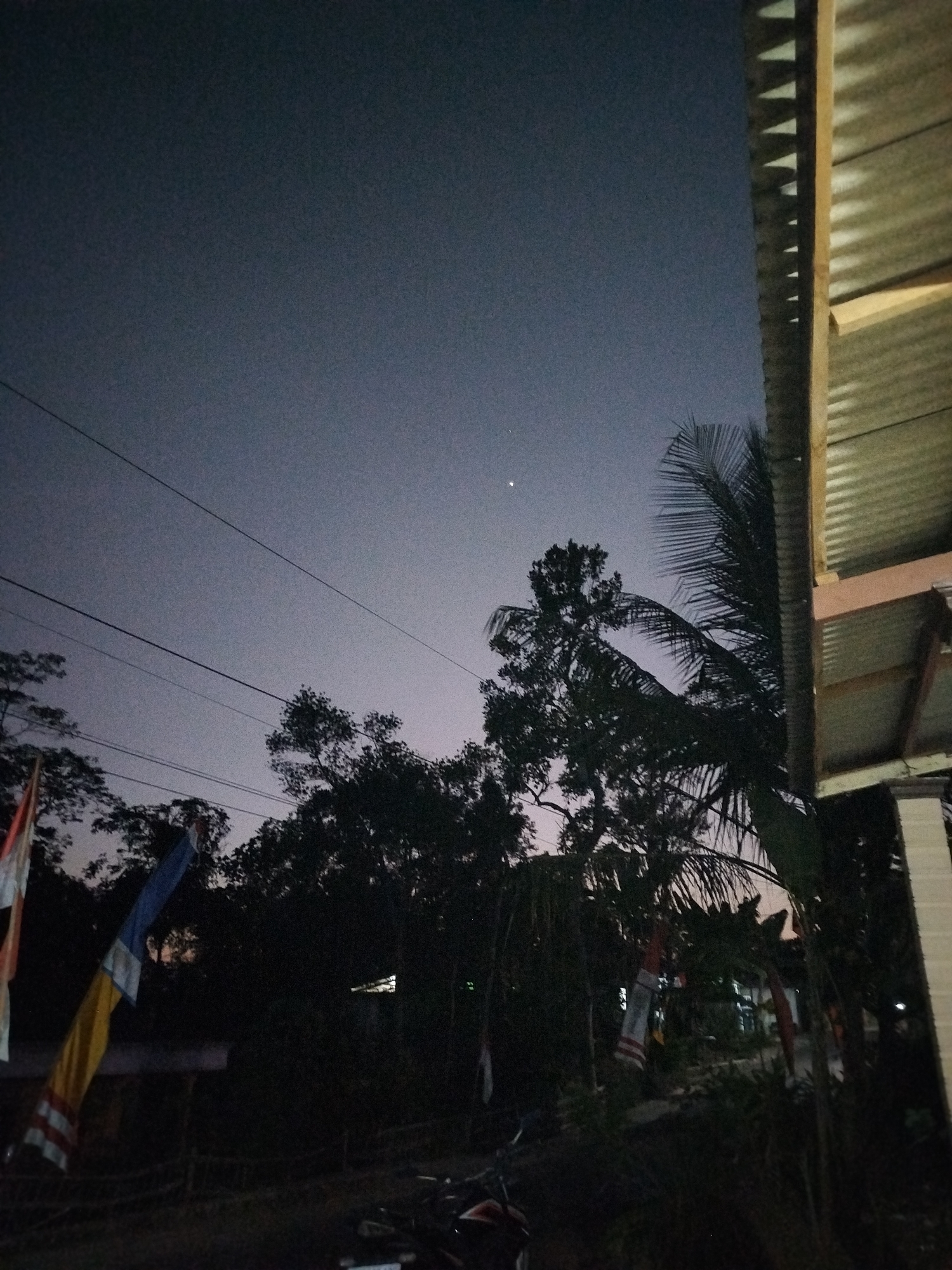 The image depicts an outdoor scene at dusk or twilight, likely in Mojogedang, Indonesia, given the provided context. The sky is a deep, dusky blue, with a single, bright point of light, possibly a star or planet, visible high in the frame. Several dark silhouettes of trees and palm fronds are scattered across the midground. Power lines are strung across the upper portion of the sky.
In the right foreground, a portion of a structure is visible, characterized by corrugated metal roofing and wooden beams. The texture of the metal is prominent, with diagonal shadows suggesting light sources from above.
In the lower portion of the image, partially obscured by darkness, there are hints of structures and objects. Flags, appearing to be red and white or red, white, and blue, are visible on the left side, along with a yellow and blue flag. The overall impression is of a rural or semi-urban setting transitioning from day to night, with elements of local culture indicated by the flags. No people are directly visible, but the presence of what appears to be a motorcycle in the bottom right corner suggests human activity in the vicinity. The lighting is low, creating deep shadows and obscuring finer details in many areas.