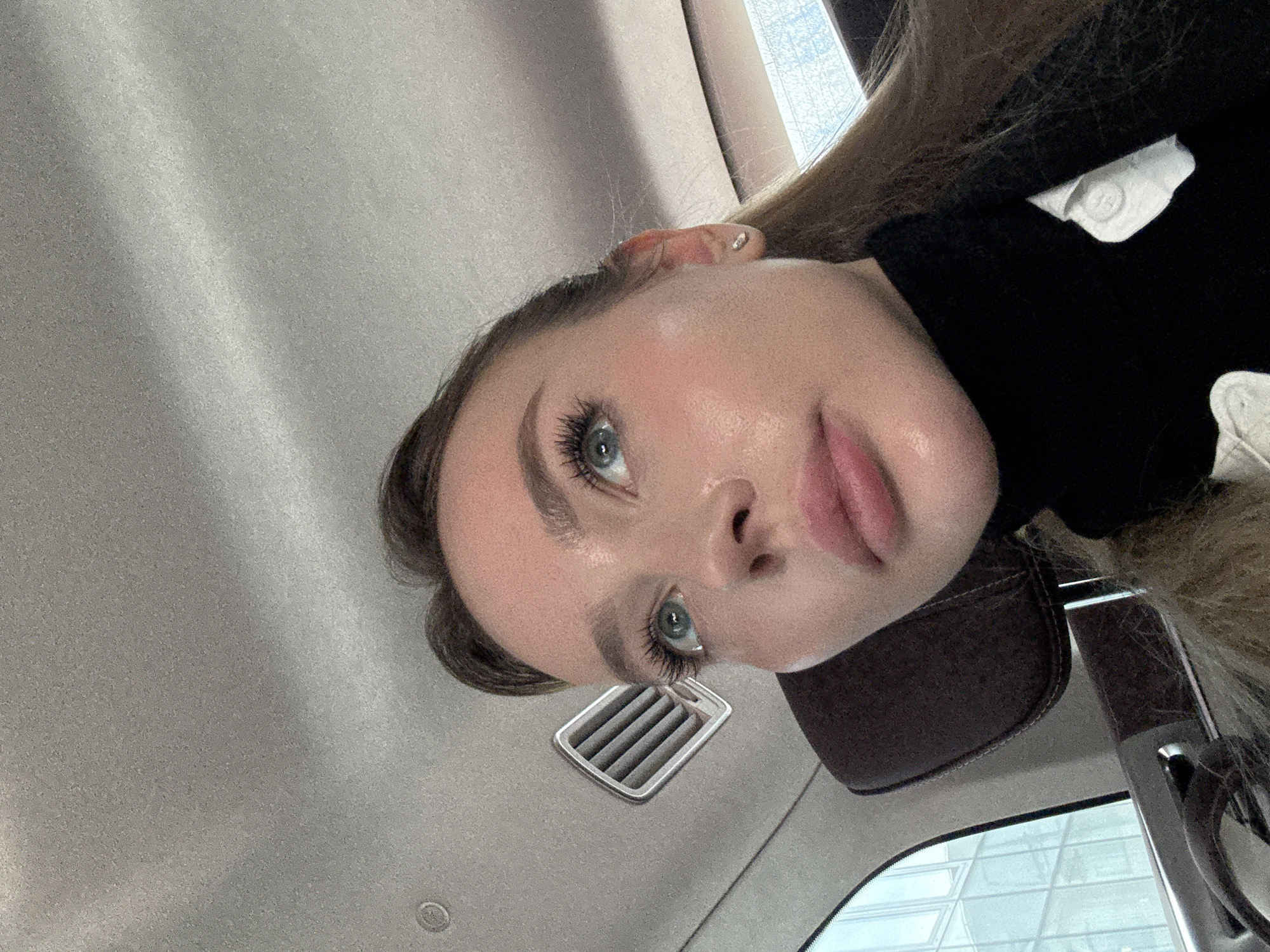 A woman is seated in a car, seemingly taking a selfie. The car's interior is visible, including the dashboard, air vents, and the headrest of the seat. Outside the car window, a building is seen reflecting light, suggesting a city setting. Given the prompt's specification, it's assumed that this location is in Moscow, Russia, though there are no explicit indicators within the image to confirm this. The woman has light skin, blue eyes, and is wearing a dark-colored top.